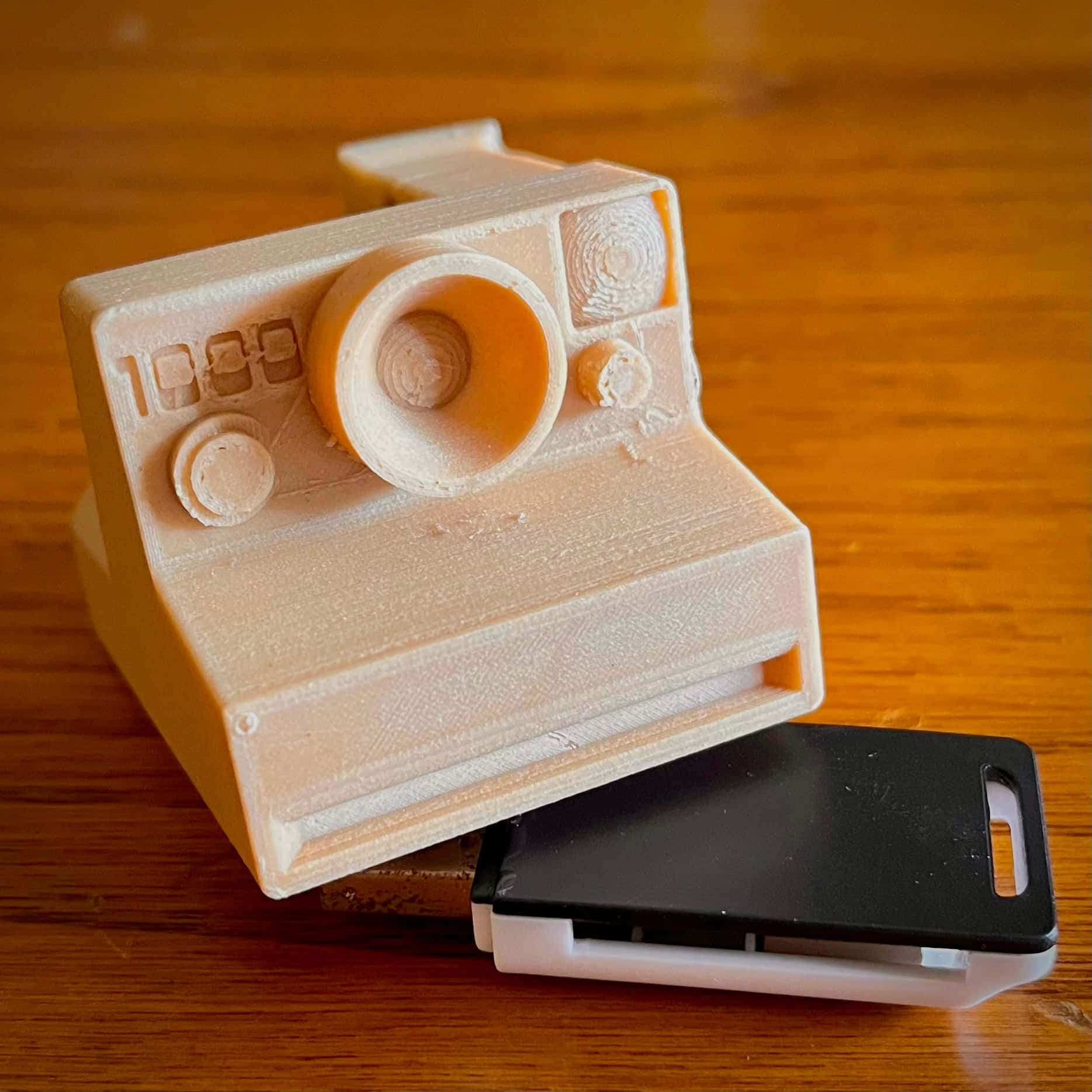  壁紙 — 一個米色3D打印即影即有相機模型放在木桌上，旁邊有一個黑色手機殼。