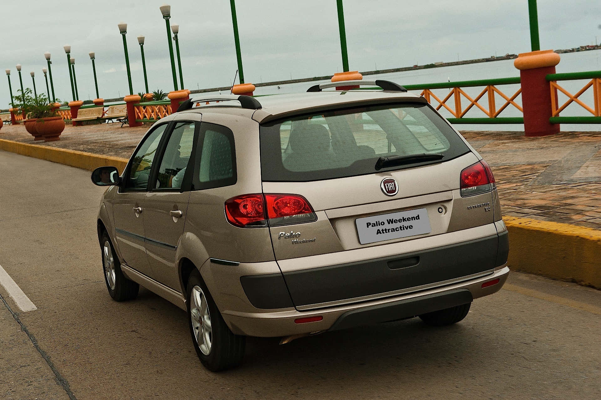 Fiat Palio 桌布 壁紙 — 一輛米色菲亞特派力奧週末吸引力旅行車停泊在海濱長廊上，周圍有色彩繽紛的欄杆和盆栽植物，天空多雲。