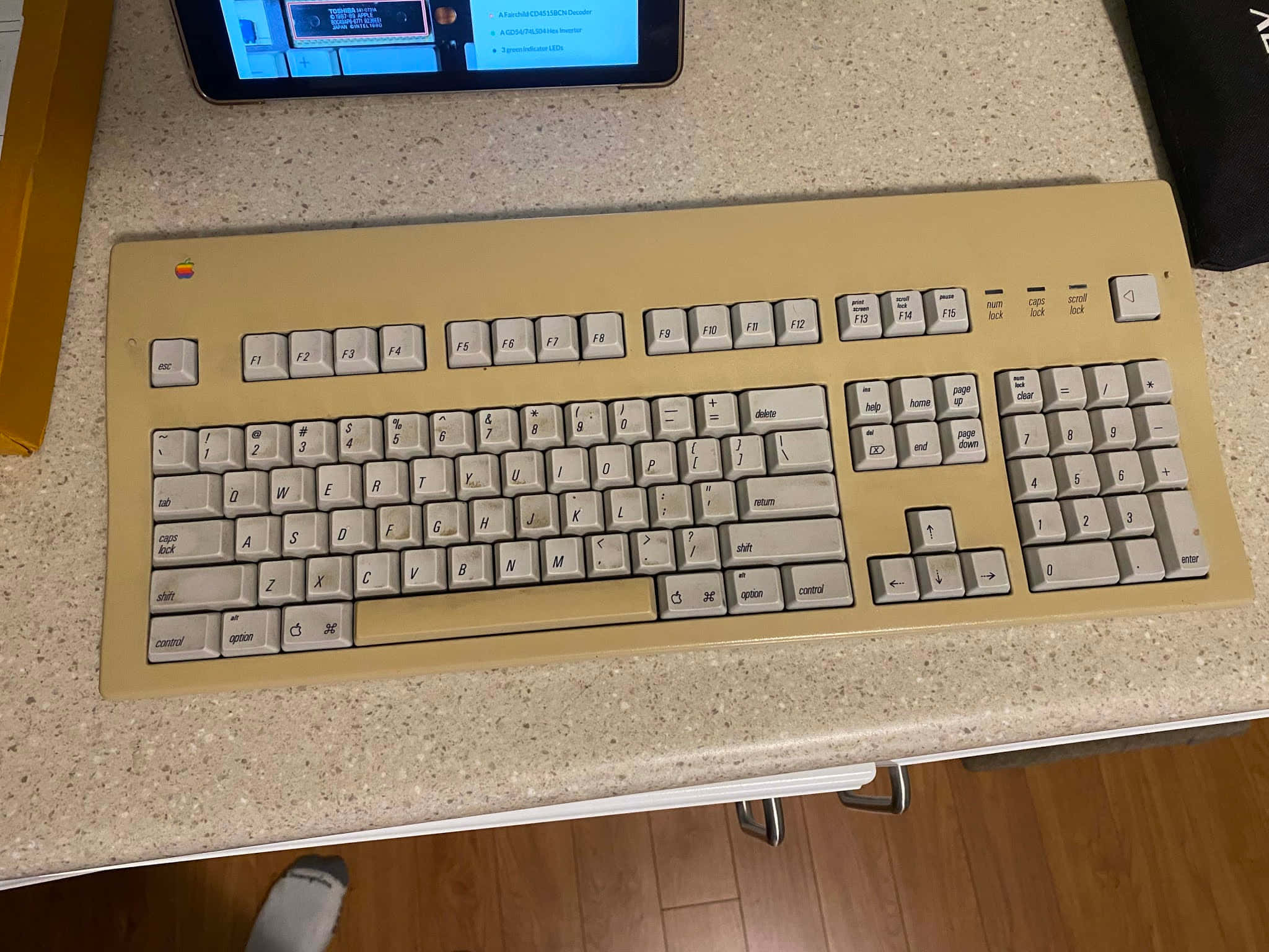 Retro Tech 桌布 壁紙 — 一個米色復古Apple鍵盤（白色按鍵）放在有斑點的檯面上，旁邊是一部顯示藍色屏幕的平板電腦。