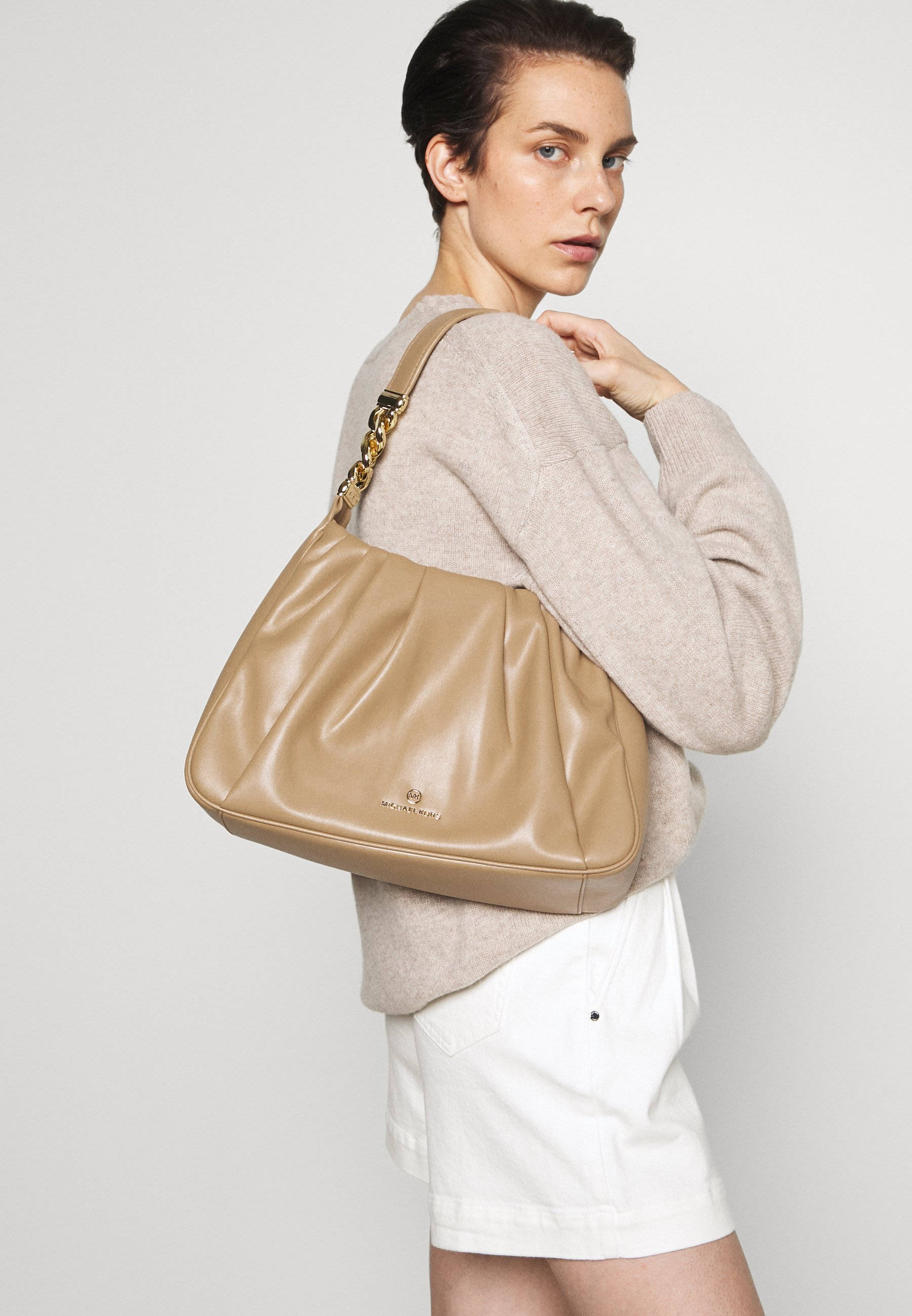 Michael Kors 米色肩背包 壁紙 — 一位身穿米色毛衣和白色短褲的女性，在純白色背景前，攜帶一個帶有金色鏈條肩帶的棕褐色皮革肩背包。