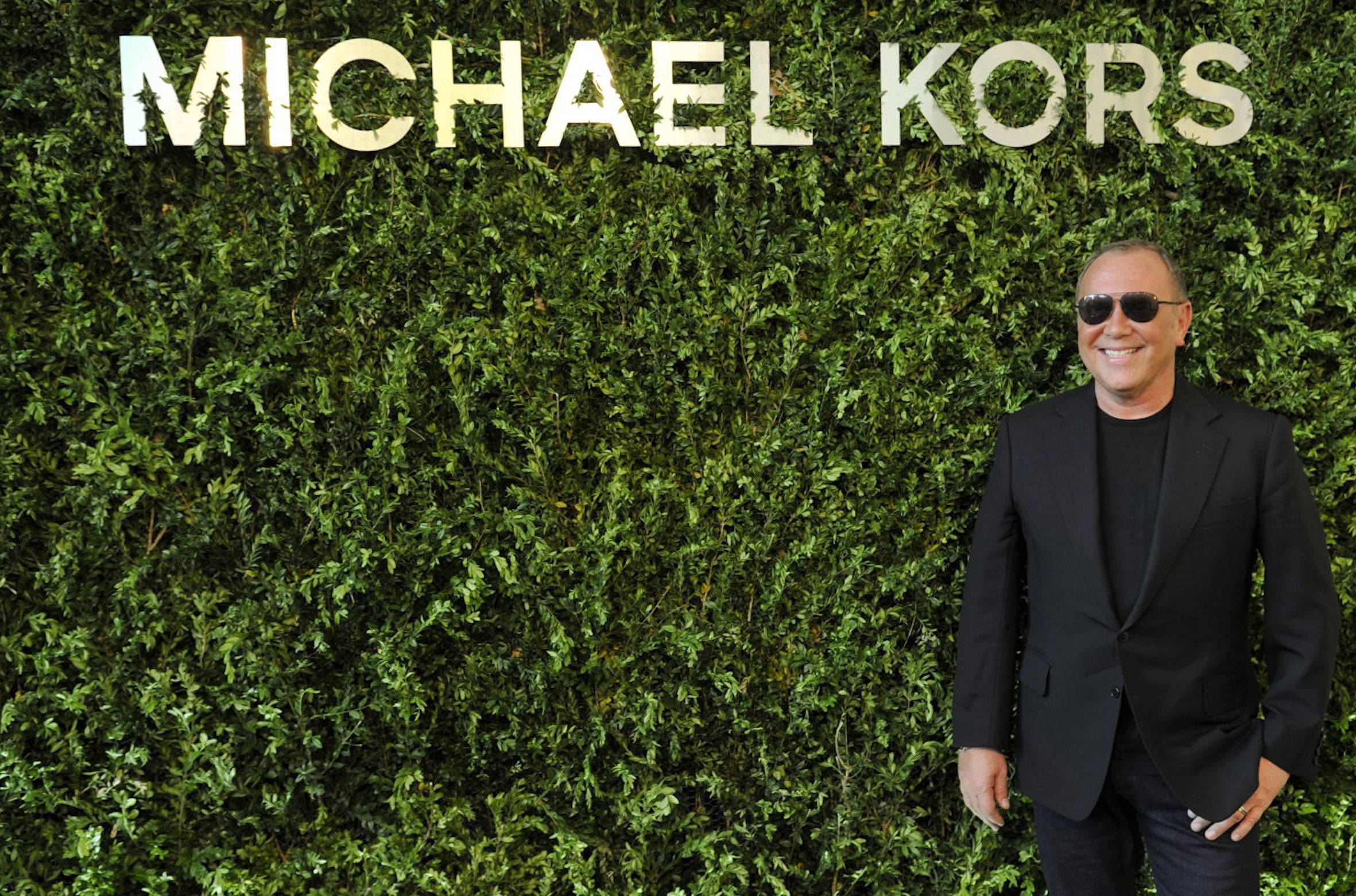 Michael Kors 全黑造型 壁紙 — 一位戴著太陽眼鏡和黑色西裝的微笑男子自信地站在茂密綠色籬笆牆前，牆上有「MICHAEL KORS」標誌。