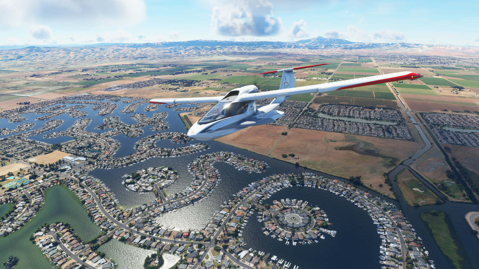 在 Microsoft Flight Simulator 中翱翔天際！ 壁紙 — 一架時尚的白色水上飛機帶有紅色機翼，在明亮的藍天下低空飛過一個擁有蜿蜒運河和遊艇碼頭的廣闊海濱社區。
