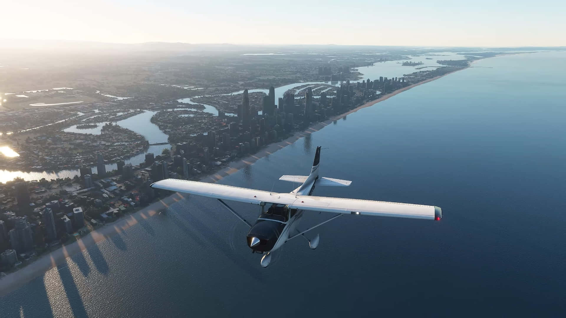 與 Microsoft Flight Simulator 一同高飛 壁紙 — 一架白色小飛機在日出時低飛過沿海城市景觀，長長的陰影在水面上延伸。