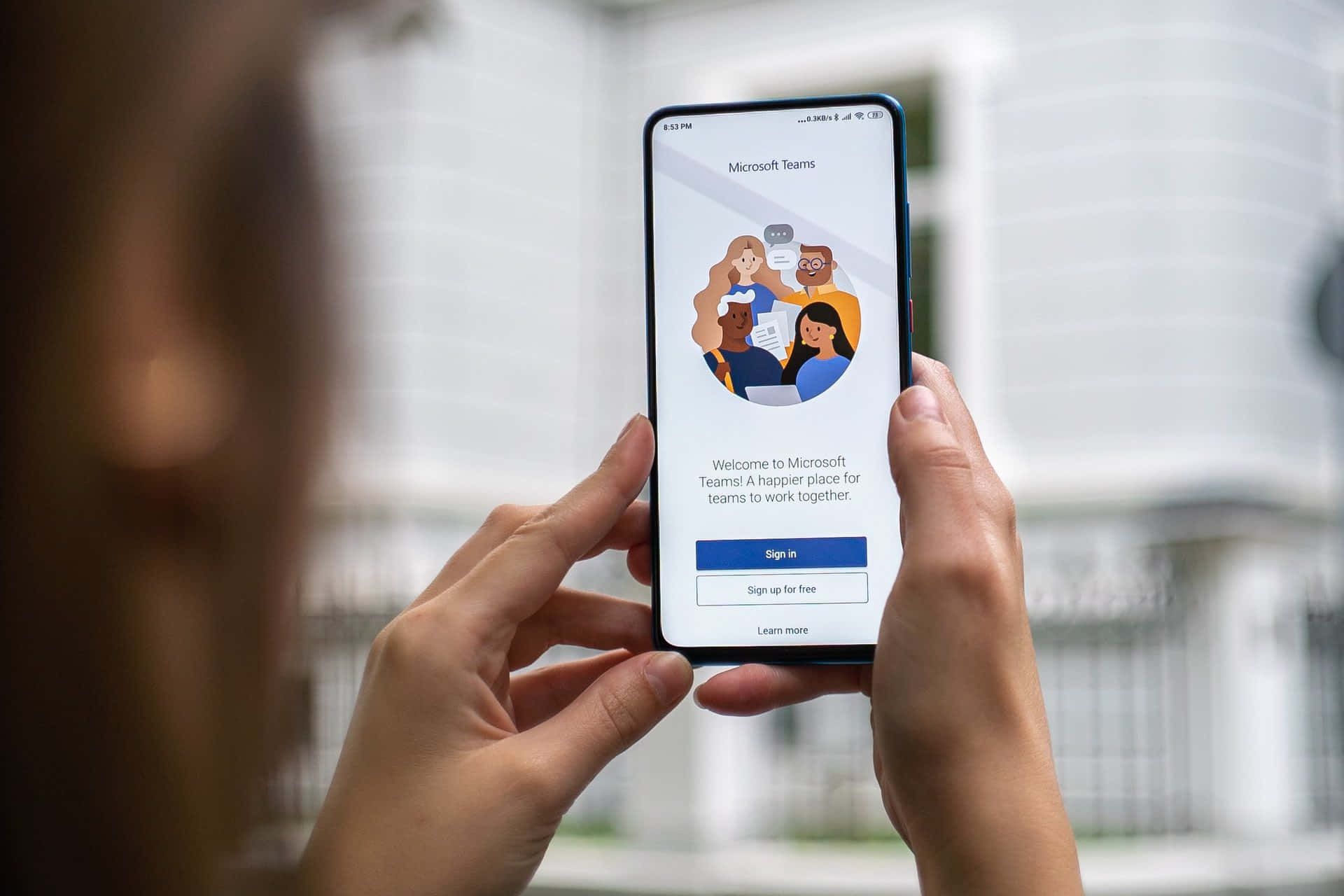 與 Microsoft Teams 一起更高效協作 壁紙 — 一個人手持智能手機，顯示Microsoft Teams歡迎畫面，上面有色彩繽紛的插圖群組和登入選項。