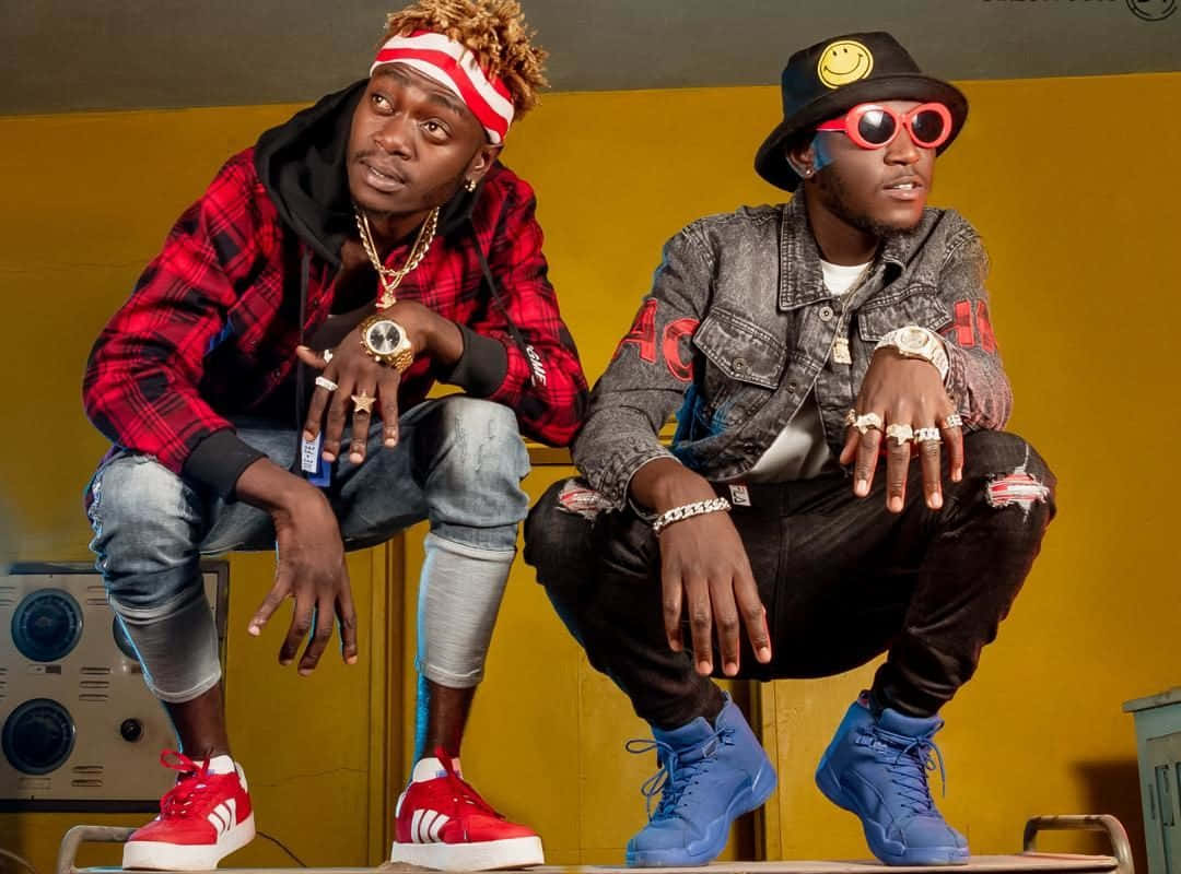 Migos 雙人組酷男孩 壁紙 — 兩位時尚男士穿著大膽街頭服飾，在充滿活力的黃色牆壁前自信擺姿勢，旁邊有復古設備。