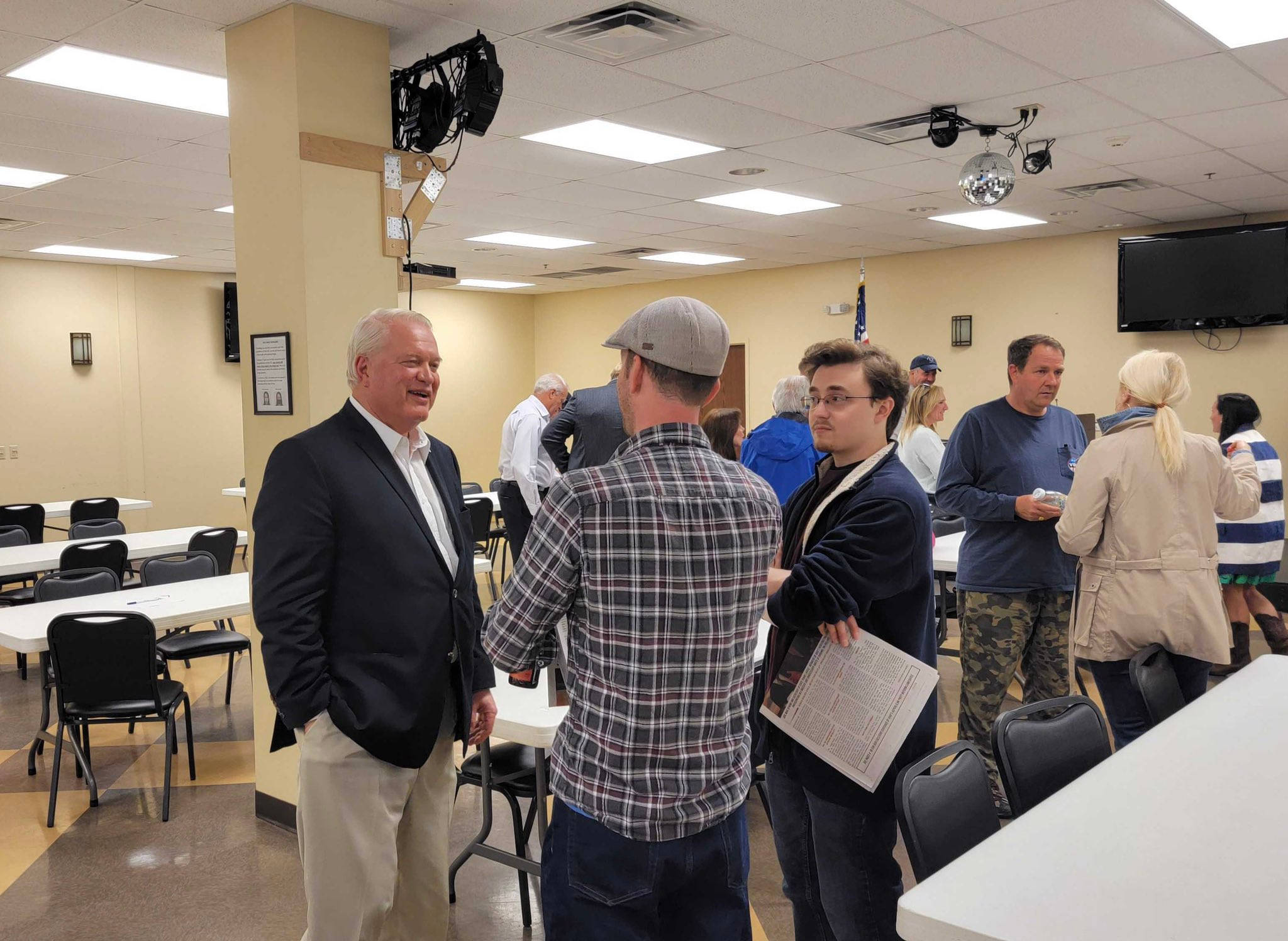 Mike Gibbons 在 East Side Republican Club 壁紙 — 人們在光線明亮的社區活動室中交流，室內有桌椅，天花板上懸掛著一顆迪斯科球。