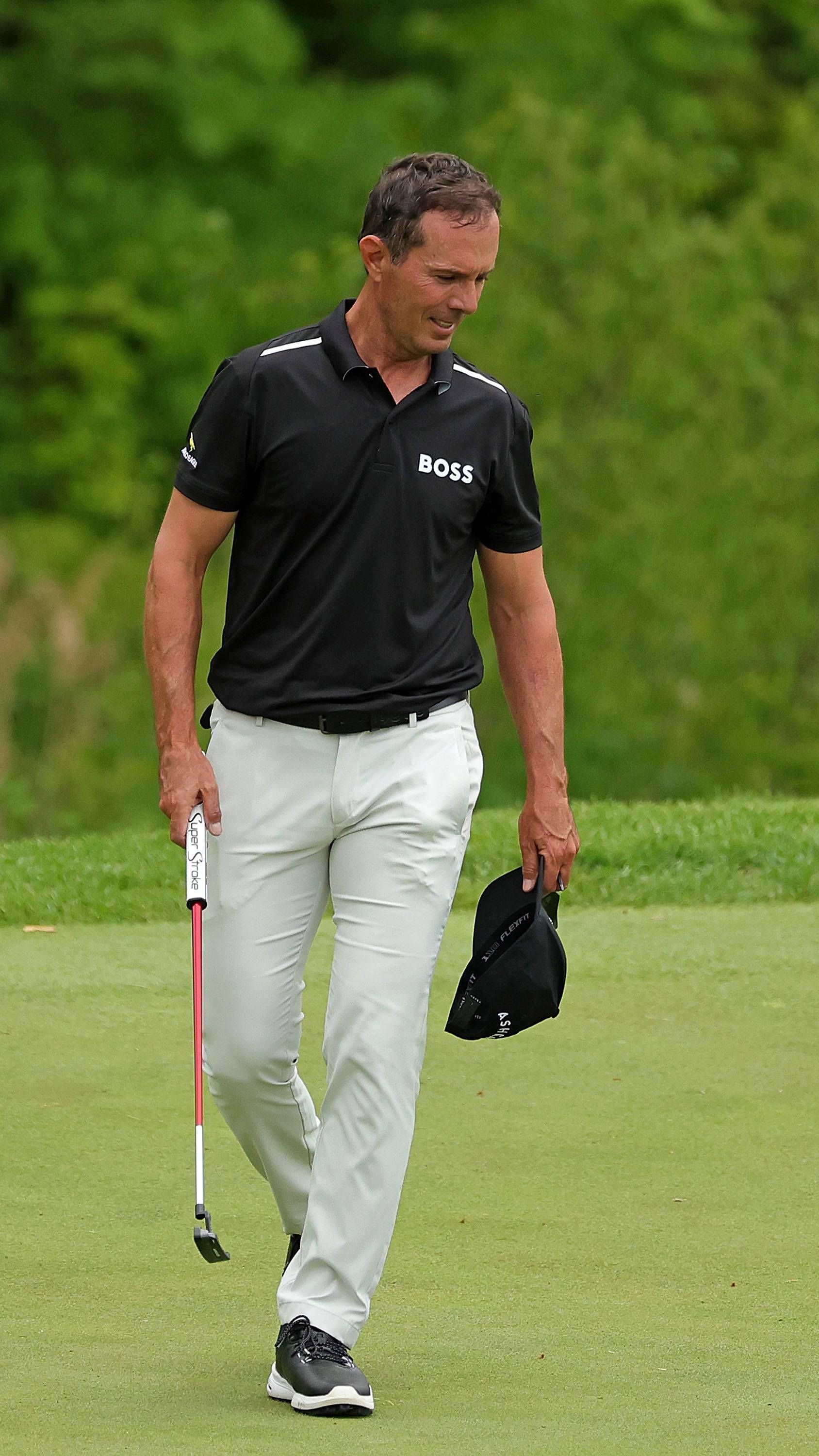 Mike Weir 球場漫步肖像 壁紙 — 一名身穿黑色Polo衫和白色長褲的高爾夫球手，手持球桿和帽子，走過翠綠的球道。