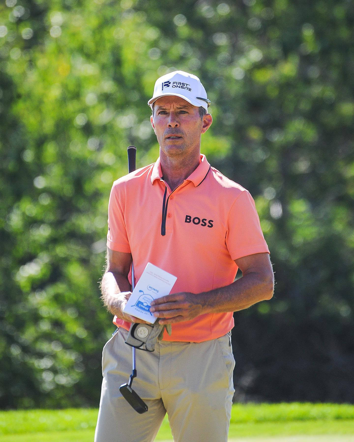 Mike Weir 穿橙色襯衫 壁紙 — 一位專注的男性高爾夫球手身穿珊瑚色Polo衫，在陽光明媚的高爾夫球場上拿著記分卡和推桿，身後是模糊的綠色樹木。