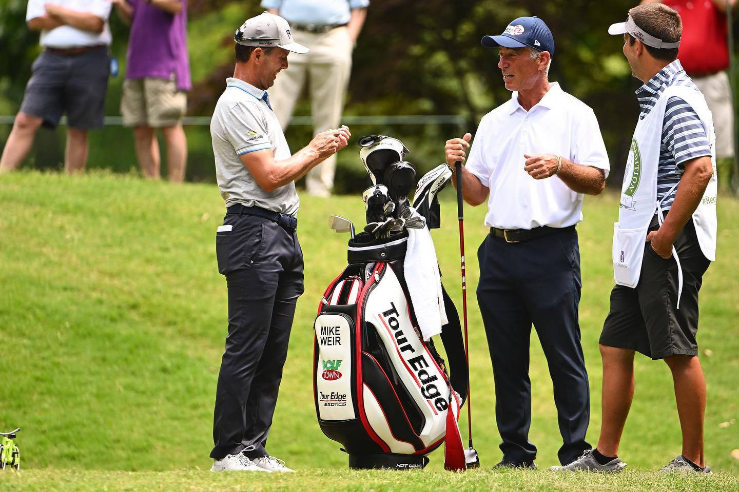 Mike Weir 與球僮同行 壁紙 — 三位高爾夫球手在陽光明媚的綠色球場上交談，他們之間有一個品牌高爾夫球袋。
