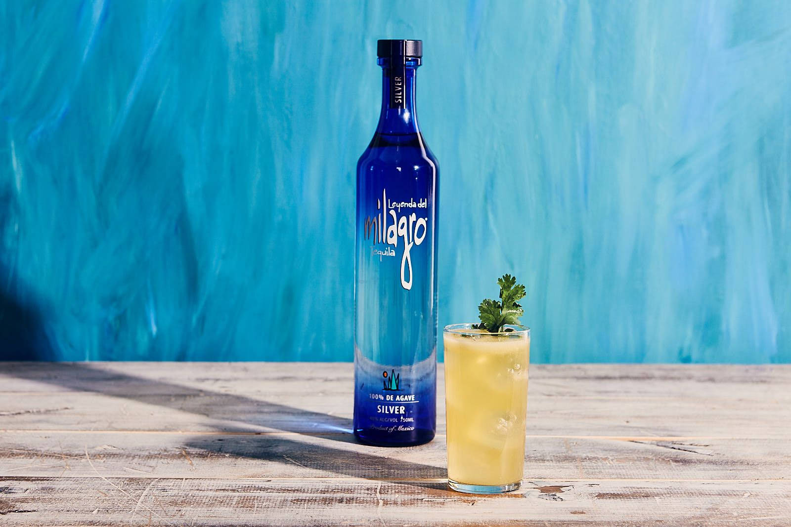 Milagro 銀色龍舌蘭酒搭配 Mexa Mula 餐飲雞尾酒 壁紙 — 一個藍色銀色龍舌蘭酒瓶站在一個黃色雞尾酒旁邊，雞尾酒上裝飾著香菜，放在一個風化的木桌上，背景是帶紋理的綠松石色。