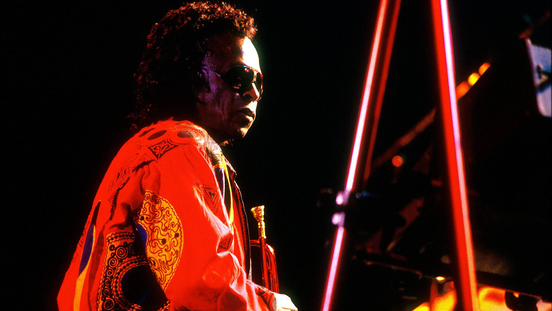 經典爵士大師 - Miles Davis 壁紙 — 一位身穿鮮豔紅色圖案外套和太陽眼鏡的音樂家在黑暗背景下，在戲劇性的紅色舞台燈光下表演。