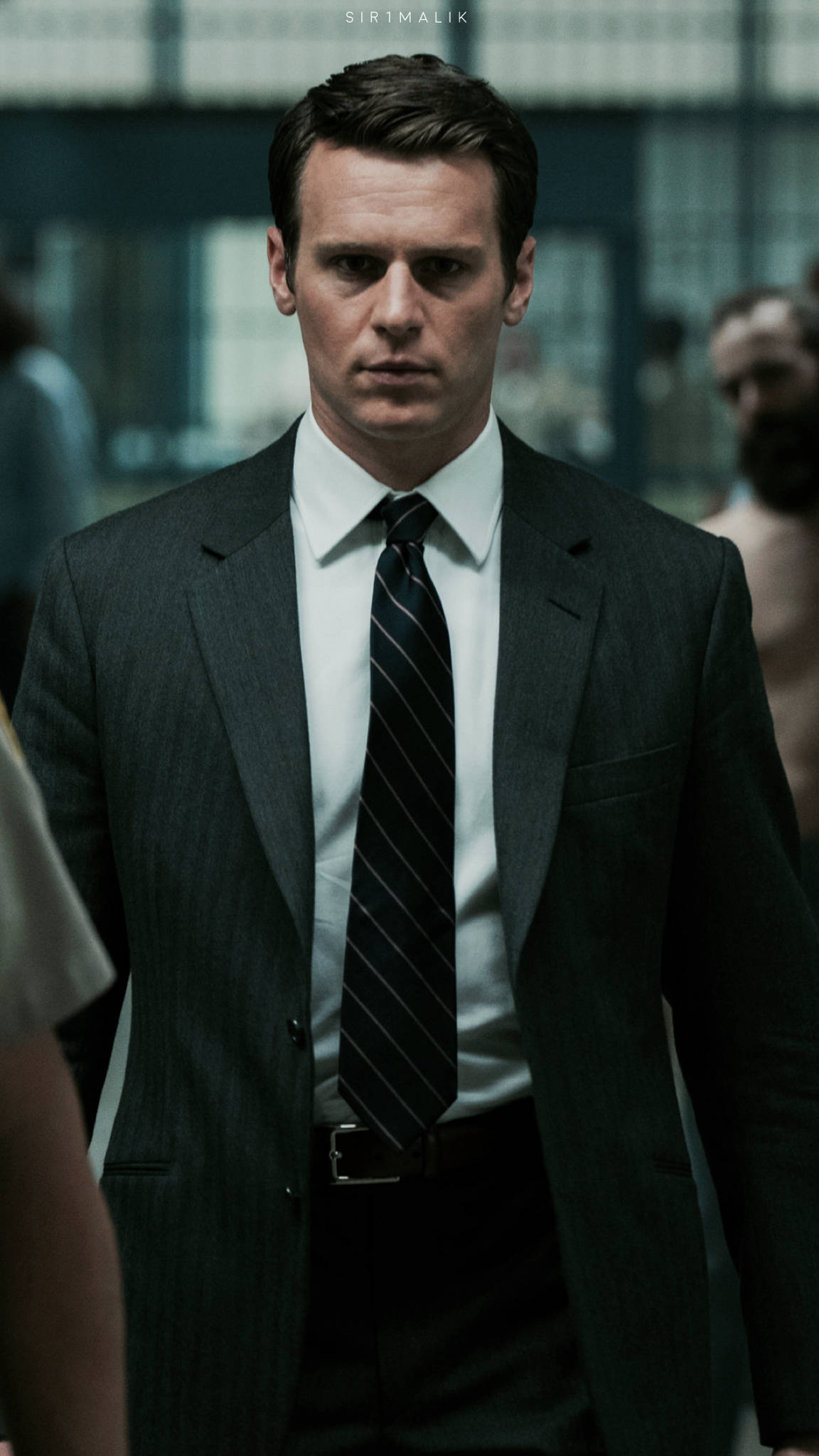 《Mindhunter》Netflix劇集演員約翰·格羅夫 壁紙 — 一名身穿深色細條紋西裝和條紋領帶的男子在光線昏暗的室內環境中專注地向前凝視。
