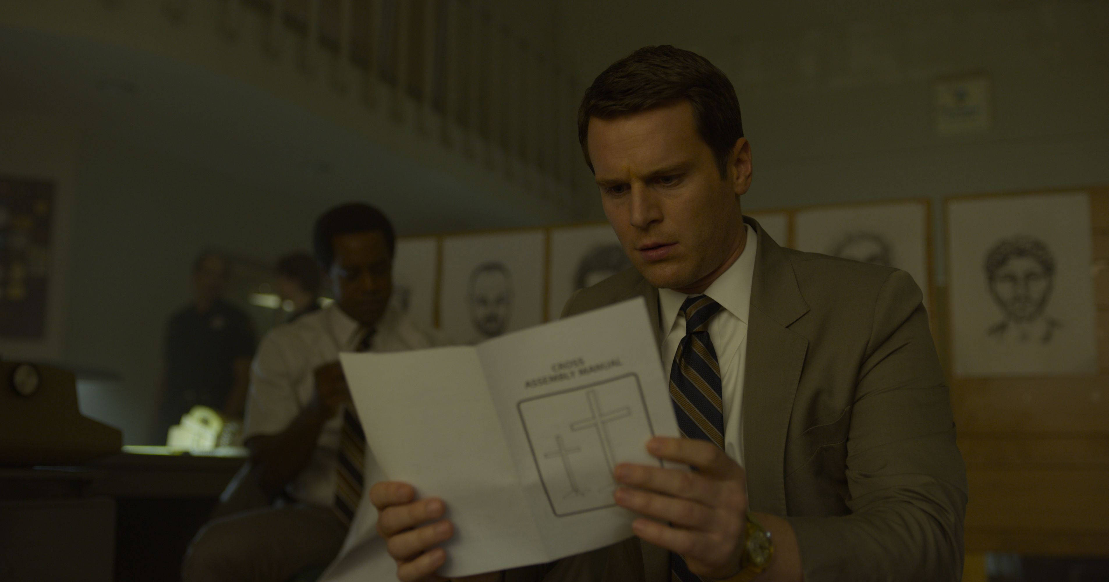 《Mindhunter》劇集主演約翰·格洛夫 壁紙 — 一名穿著西裝的男子手持帶有十字架草圖的葬禮程序單，其他人則坐在光線昏暗的房間裡，牆上掛著帶框的肖像。