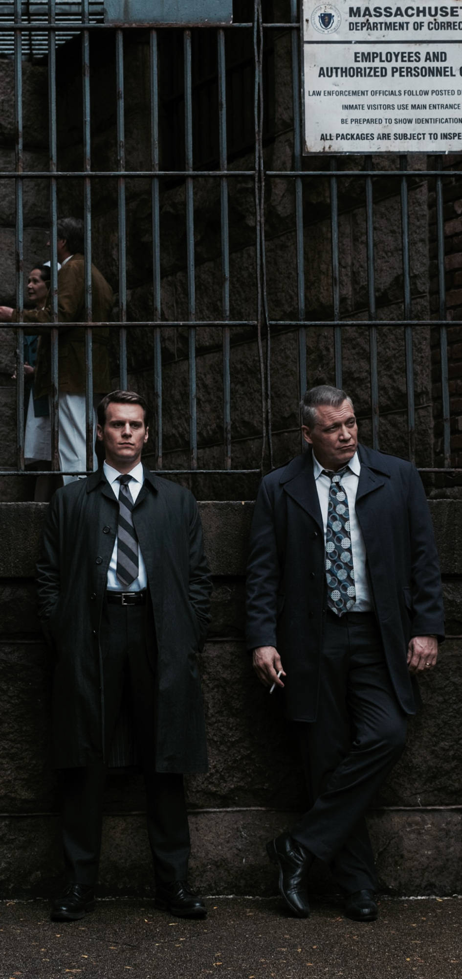 《Mindhunter》霍爾登·福特與比爾·坦奇 壁紙 — 兩名身穿深色外套的男子站在麻省監獄外的石牆旁，監獄設有鐵閘和標牌。