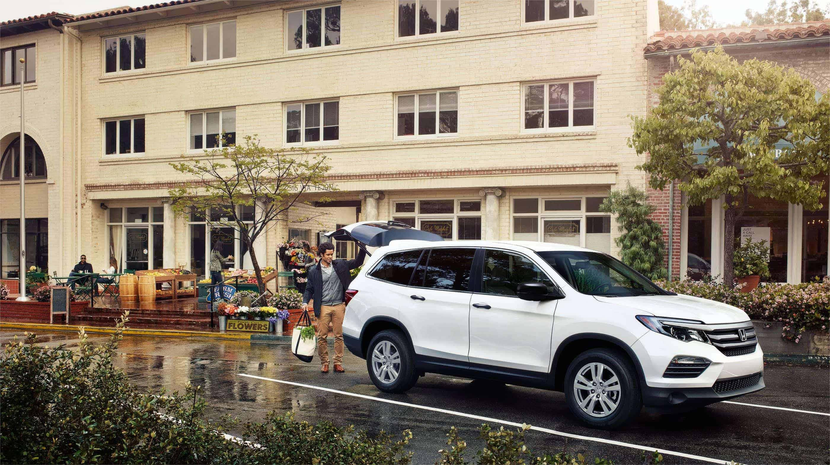 Honda Pilot 桌布 壁紙 — 一名男子站在一輛白色SUV旁邊，尾門打開，位於潮濕的停車場，附近有花店和米色建築物。
