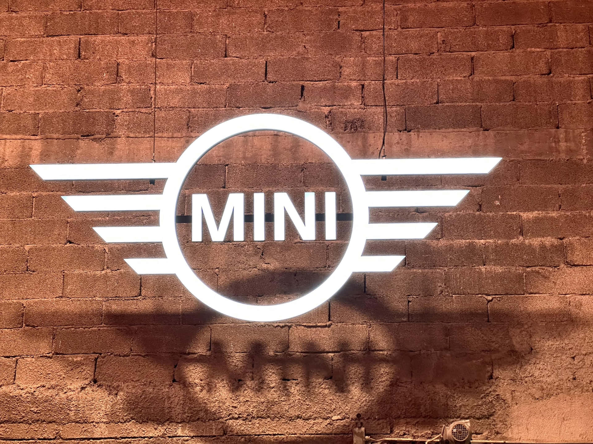 Mini Cooper標誌投影於磚牆 壁紙 — 一個發光的白色MINI標誌，帶有翼形徽章，在紋理紅磚牆上投射出清晰的陰影。