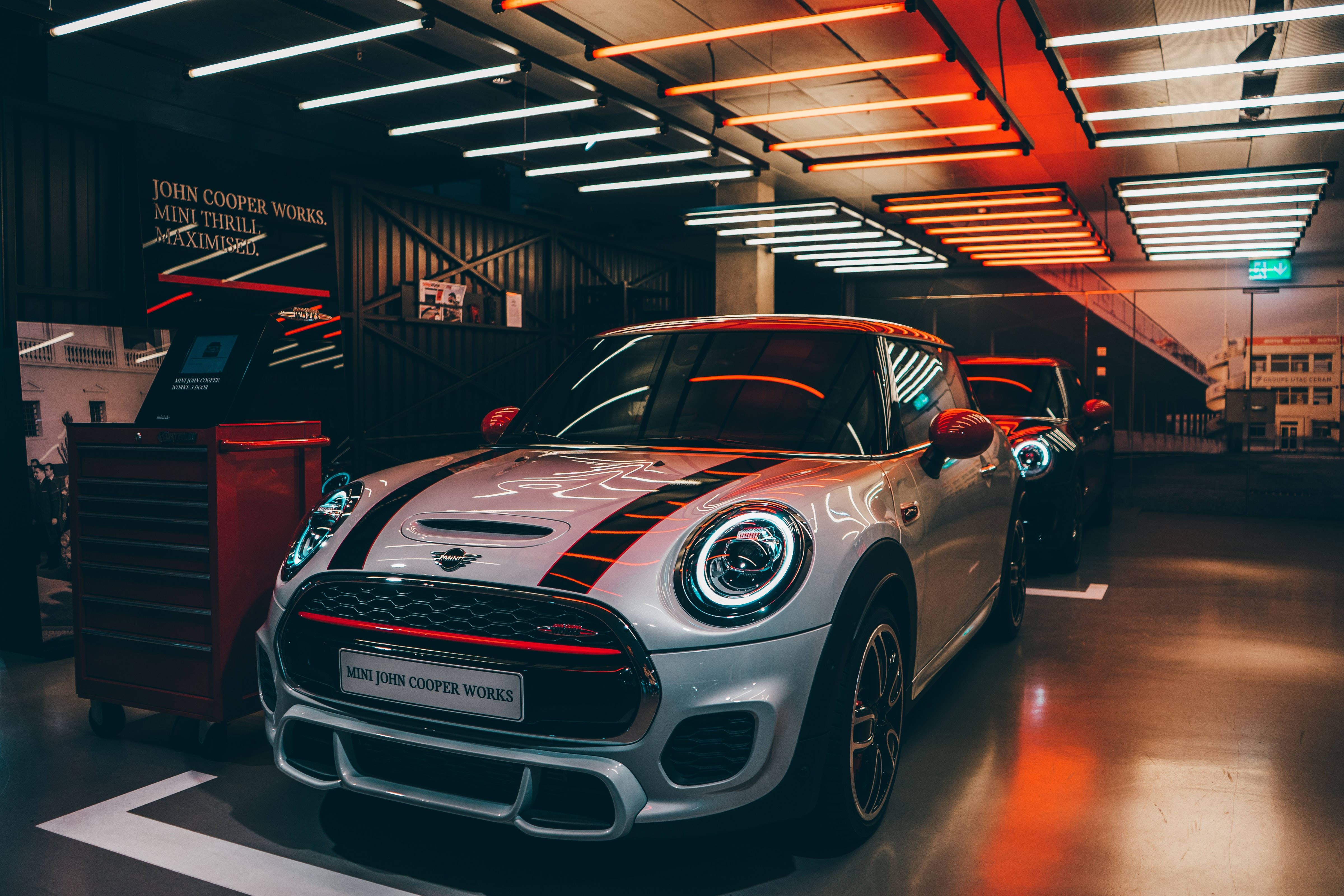 Mini 車款於霓虹美學停車場 壁紙 — 一輛時尚的銀色MINI John Cooper Works在現代車庫中，在充滿活力的橙色和白色天花燈下閃閃發光。