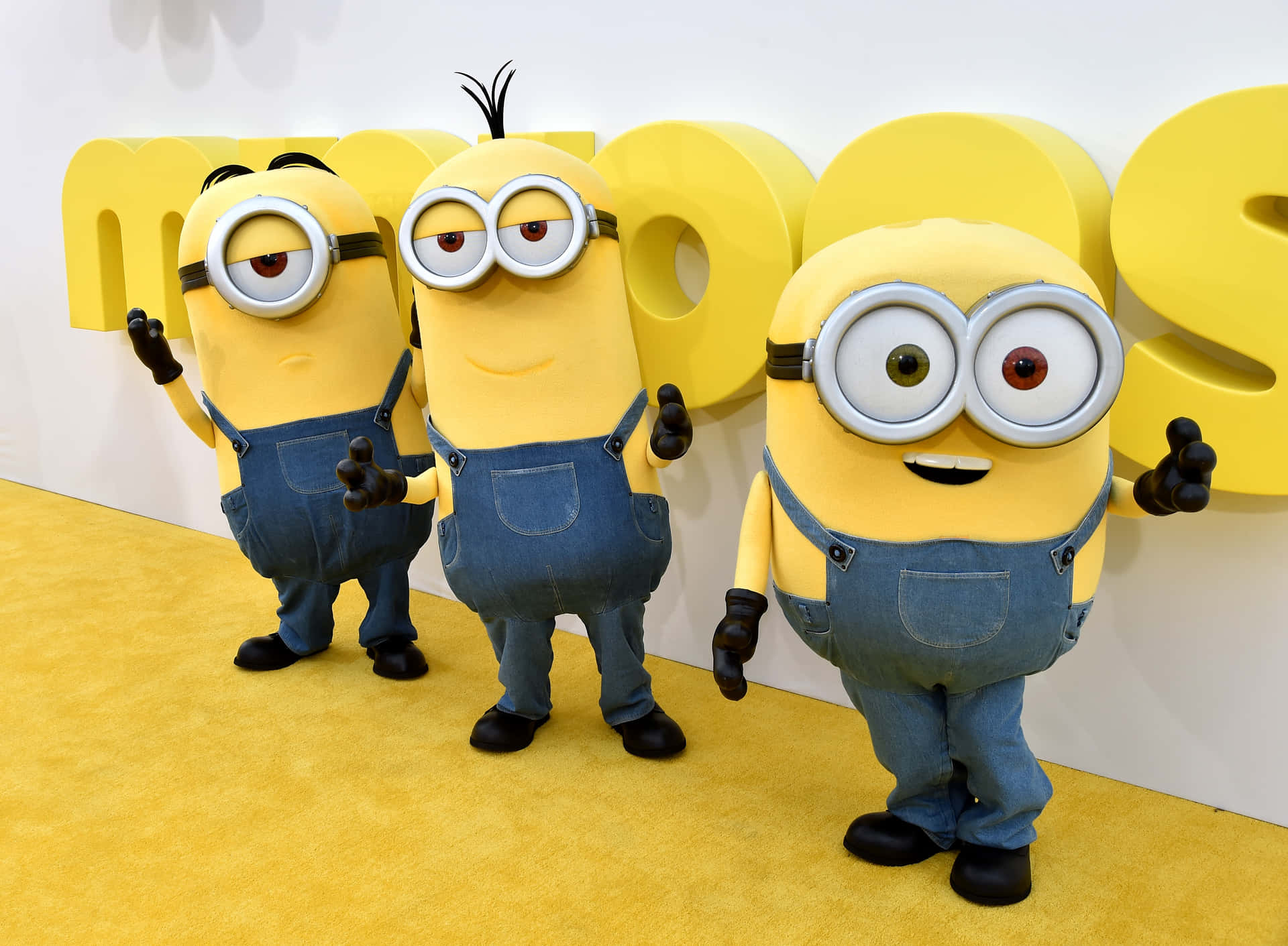 三個穿著藍色工裝褲的Minions在活動中在黃色地毯上擺姿勢。