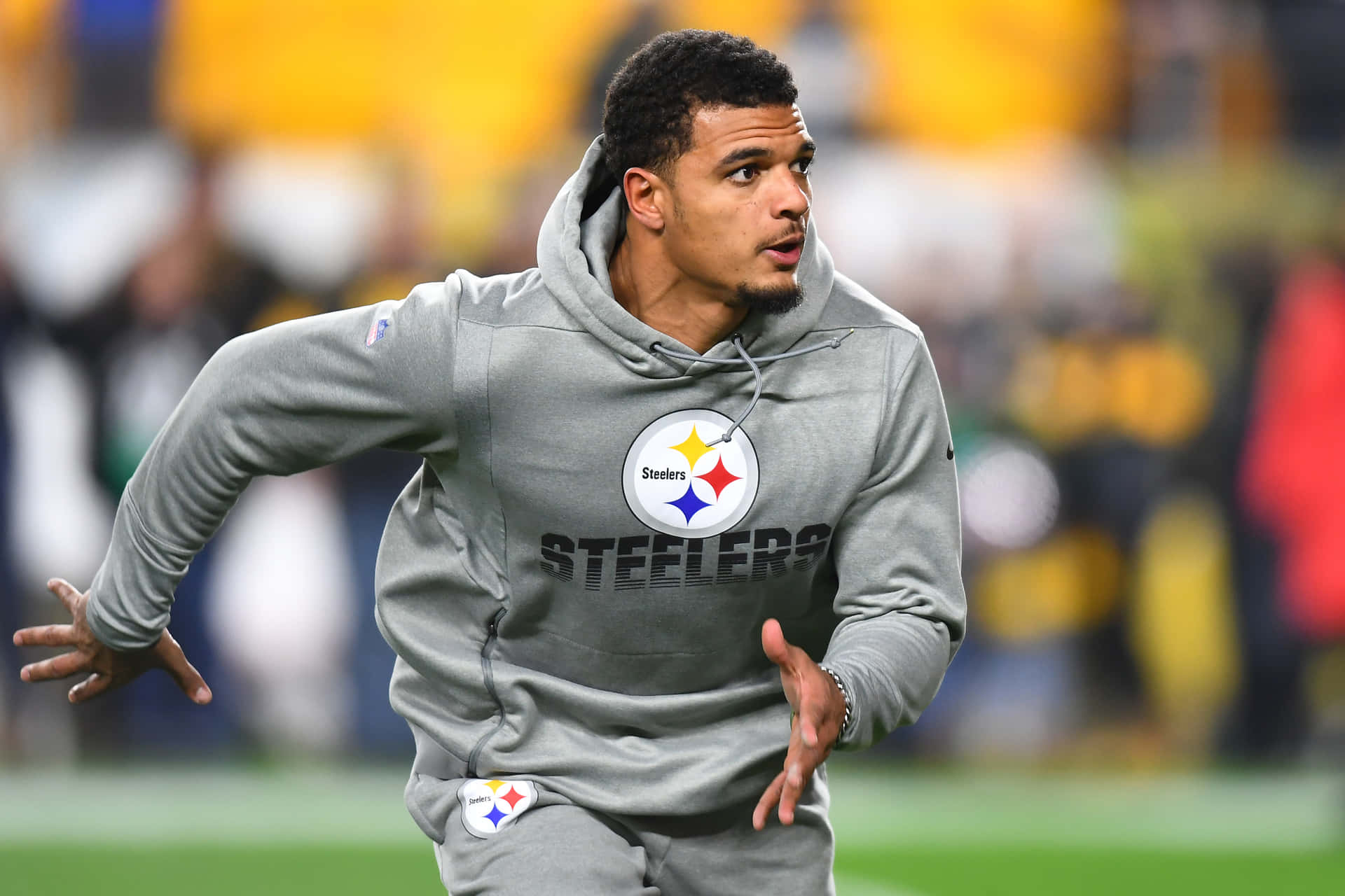 Minkah Fitzpatrick 匹茲堡鋼人隊暖身連帽衫 壁紙 — 一名身穿灰色連帽衫的匹茲堡鋼人隊球員在綠色球場上衝刺，背景是模糊的體育場人群。