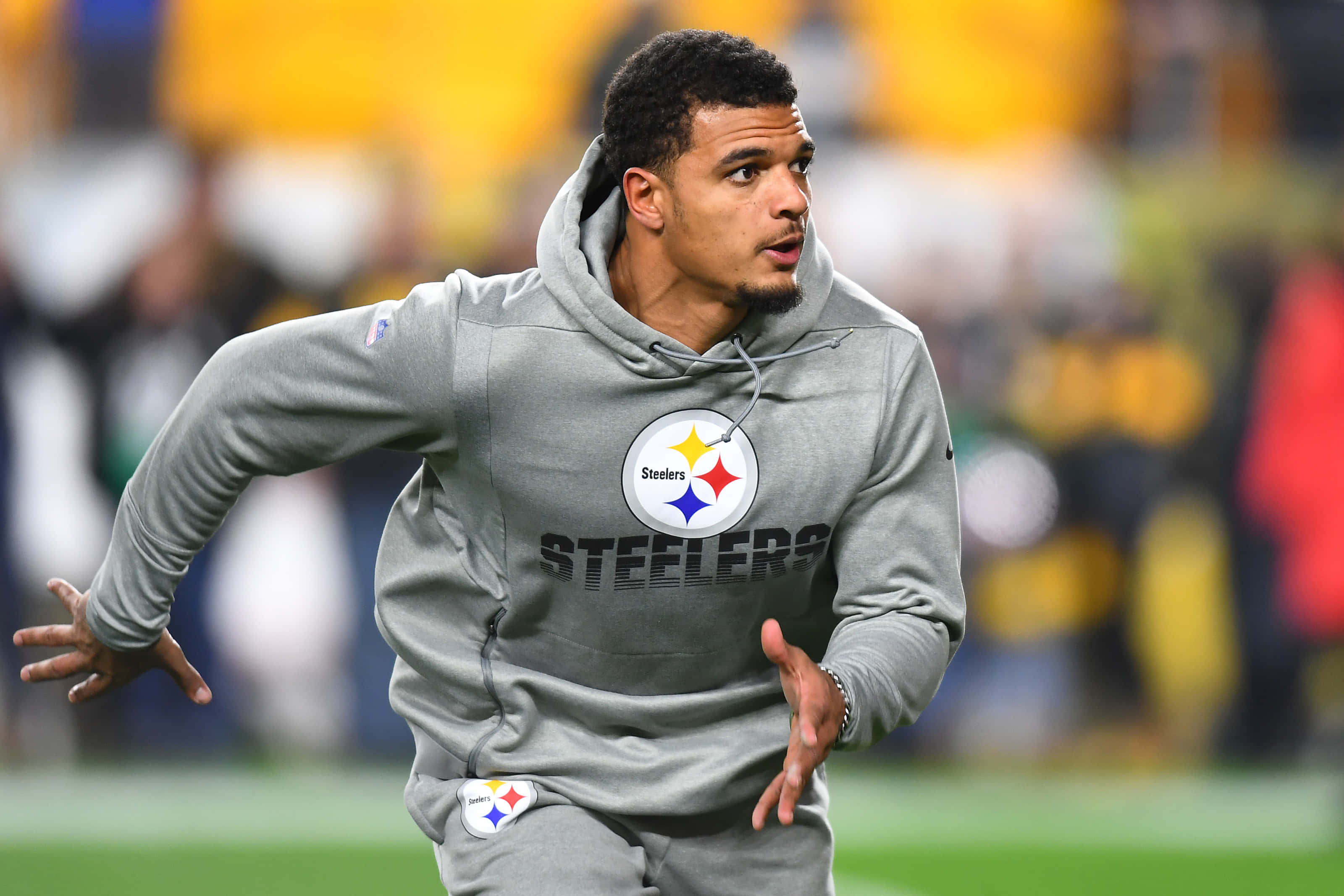 Minkah Fitzpatrick 匹茲堡鋼人隊暖身連帽衫 壁紙 — 一名身穿灰色連帽衫的匹茲堡鋼人隊球員在綠色球場上衝刺，背景是模糊的體育場人群。