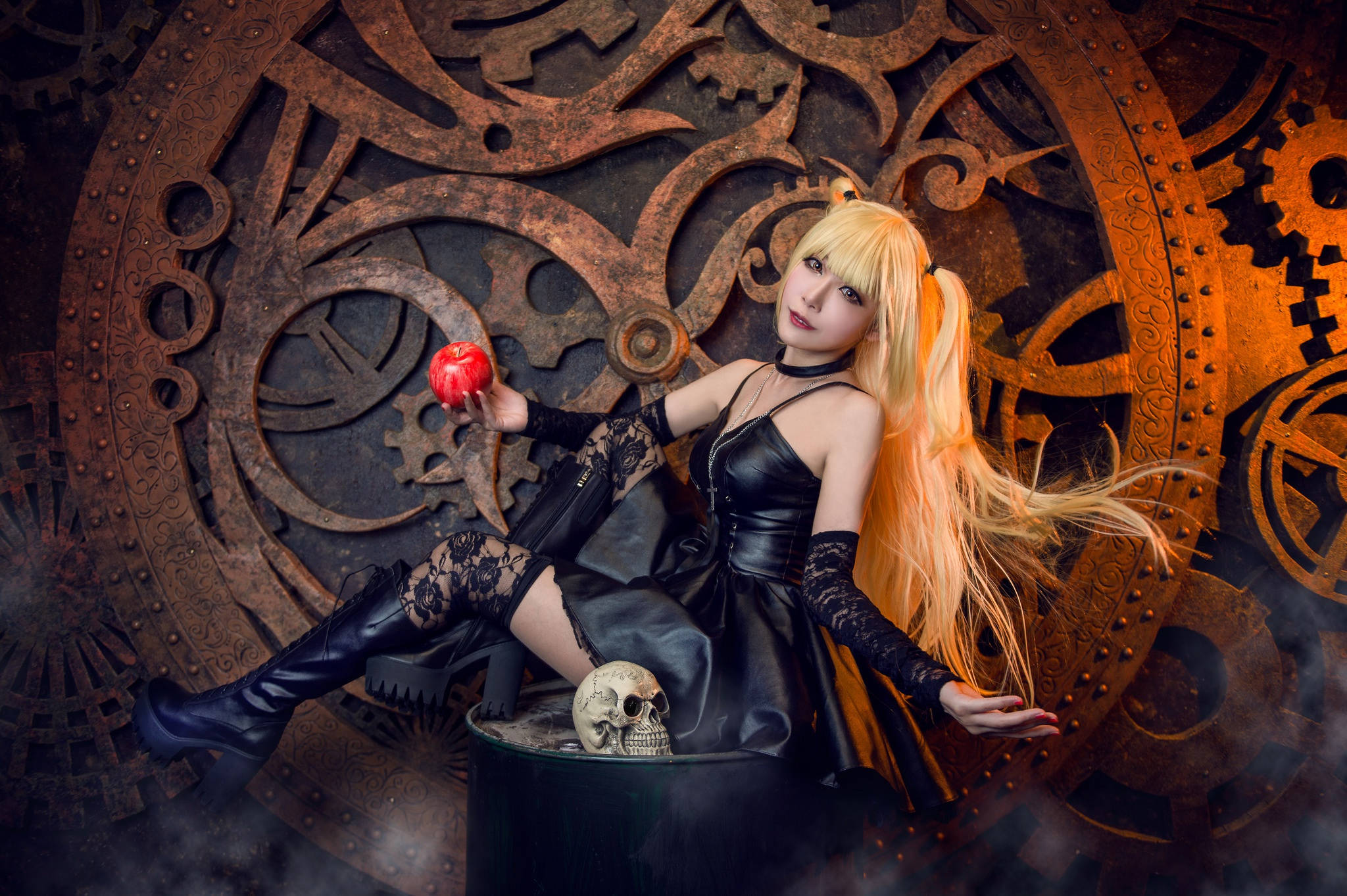 Misa Amane 全身Cosplay 壁紙 — 一位金髮女子身穿黑色皮革，手持紅蘋果，背景是華麗的鏽蝕齒輪和一個頭骨。