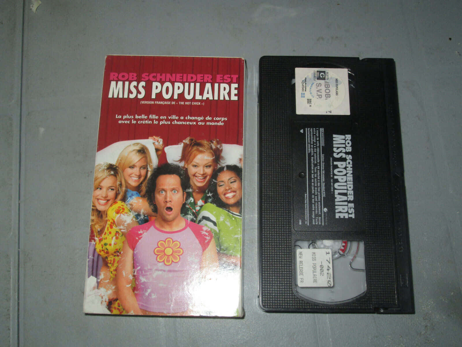 羅伯·施奈德《Miss Populaire》專輯封面 壁紙 — 《Rob Schneider est Miss Populaire》的VHS錄影帶及其封套並排放在灰色表面上。