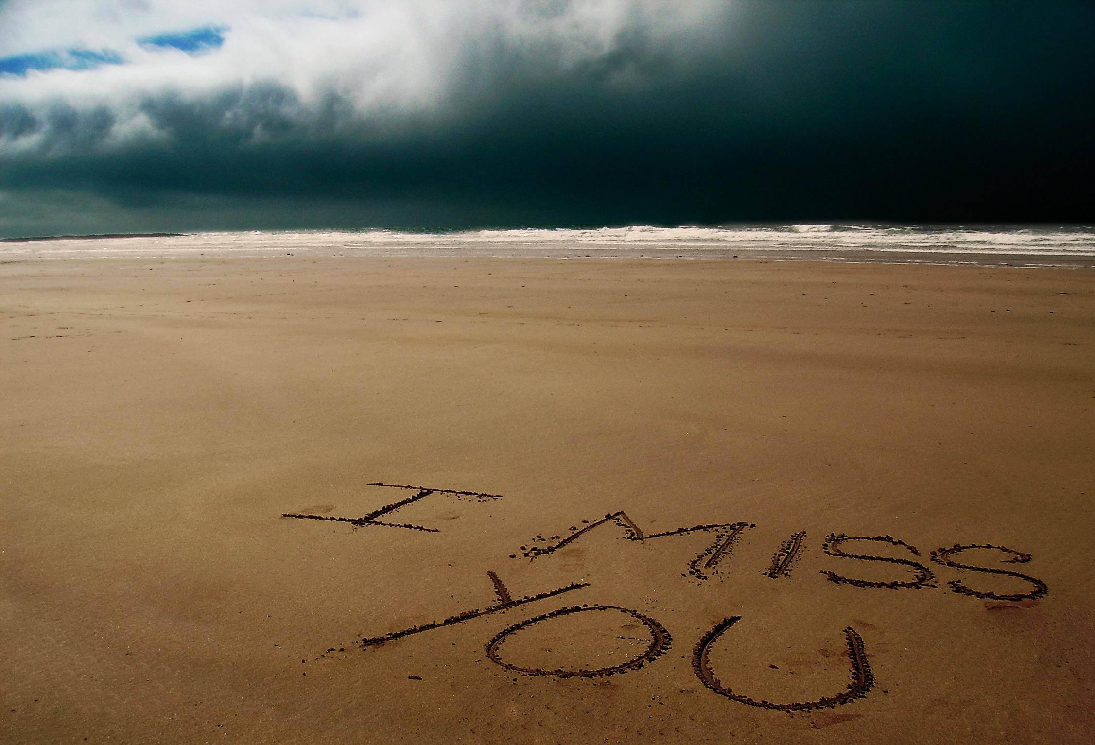 《Missing You》沙灘文字 壁紙 — 「I MISS YOU」呢幾個字刻喺一個廣闊沙灘嘅濕沙上，上面係一片逼近嘅黑暗暴風雲。