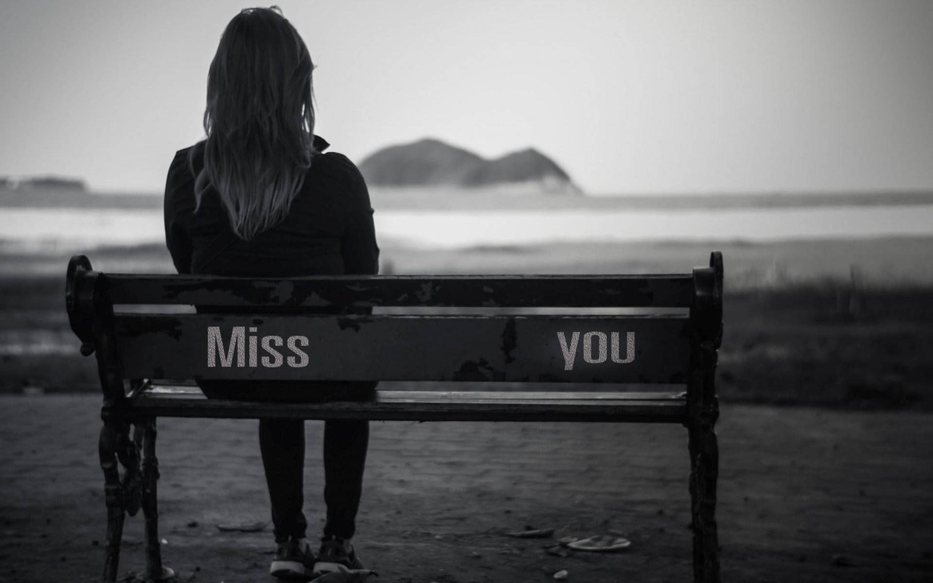 《Missing You》獨坐長椅 壁紙 — 一個孤獨的人坐在水邊一張飽經風霜的長椅上，椅背上以單色繪有「Miss you」字樣。