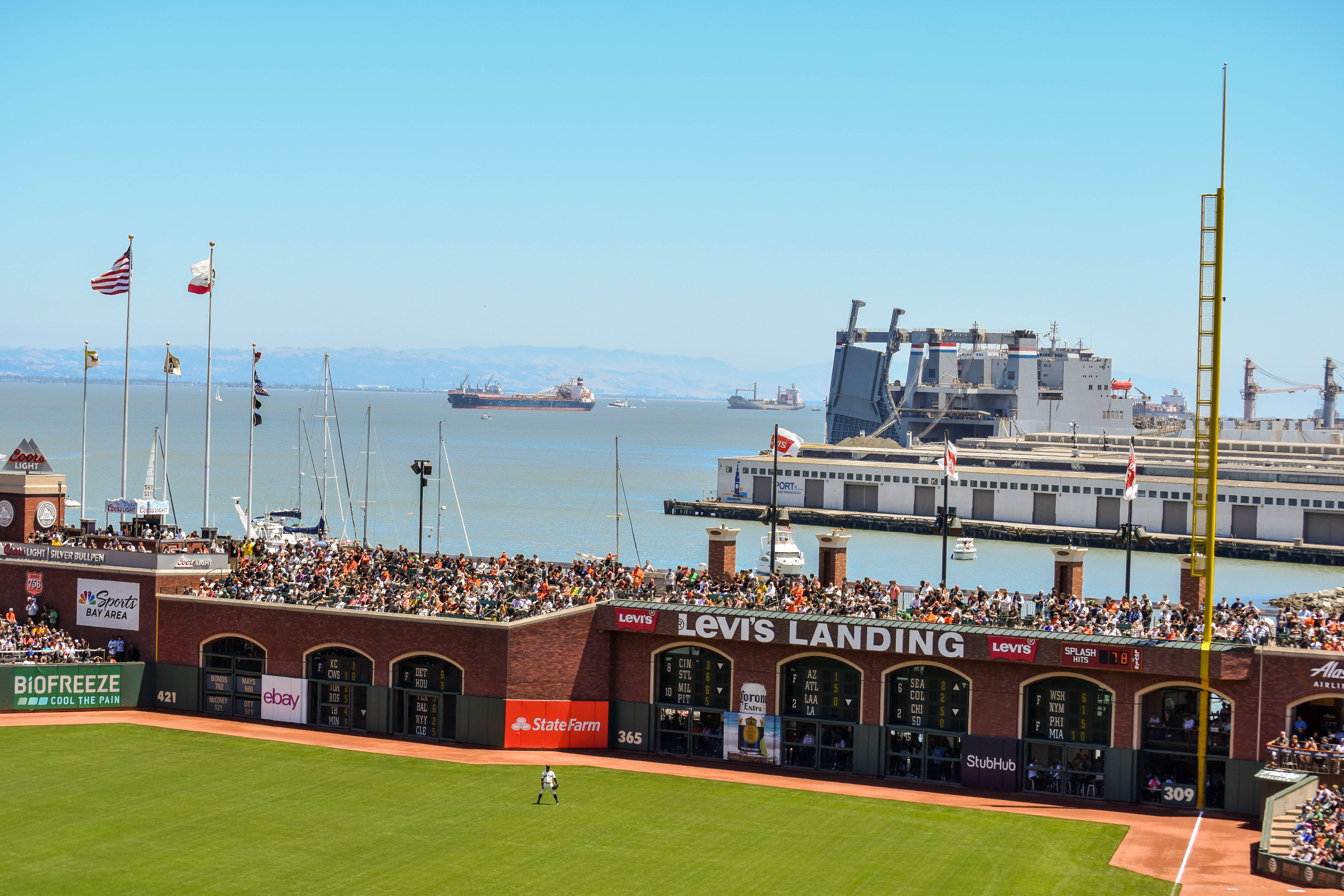 MLB 棒球賽南灣 壁紙 — 一個陽光明媚的棒球場擠滿了球迷，俯瞰著一個有船隻和大型造船廠的港口，天空湛藍。