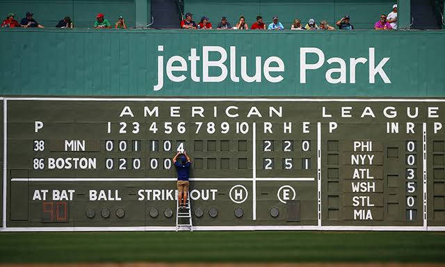 MLB比分：波士頓對明尼蘇達 壁紙 — 一名工人喺梯上更新jetBlue Park嘅記分板，期間有棒球比賽進行，觀眾喺看台觀看。