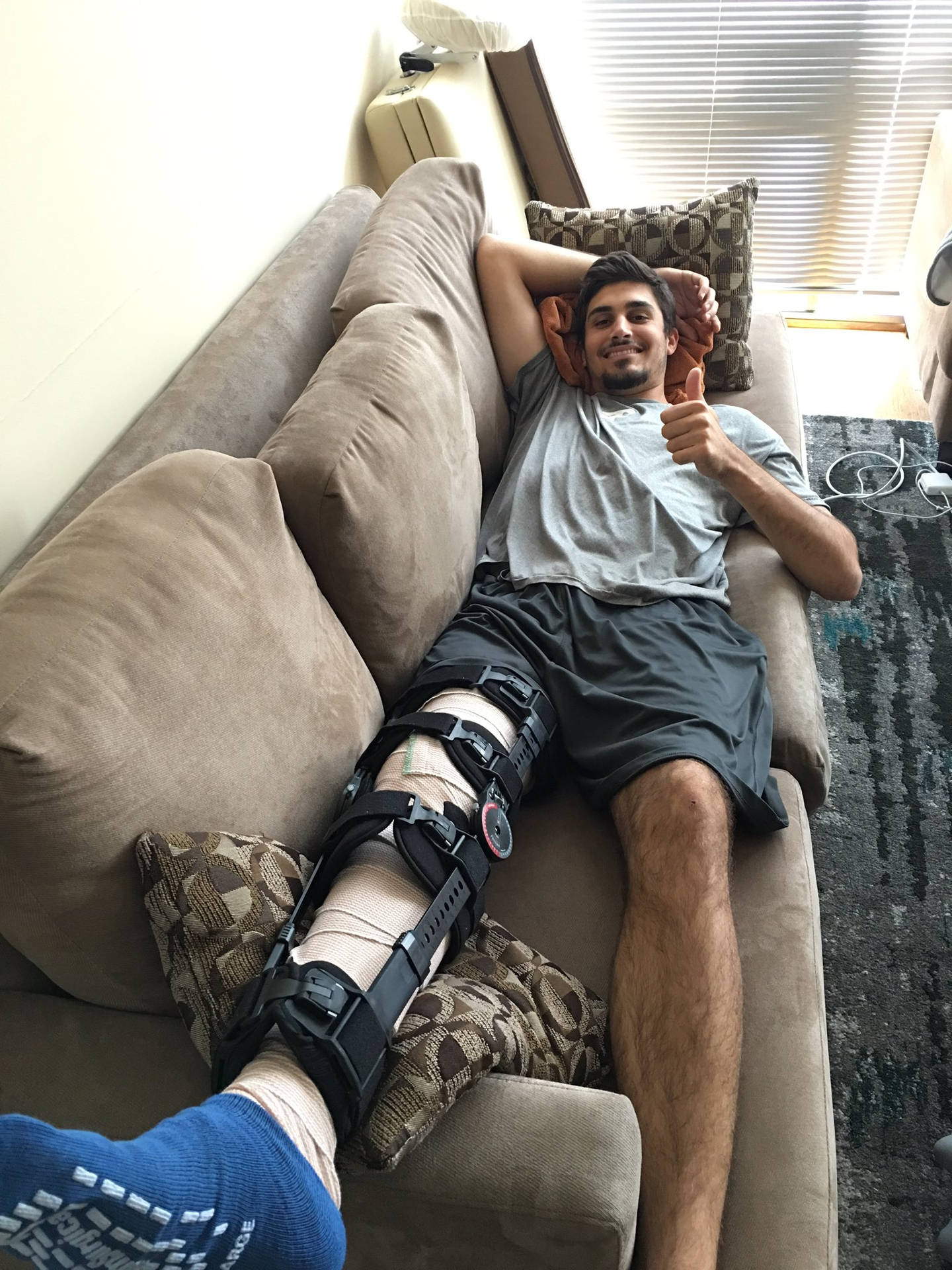 MLB球員Zach Eflin腿部受傷 壁紙 — 一位微笑的男子，腿上打著石膏並穿著靴子，在米色沙發上放鬆並豎起大拇指。