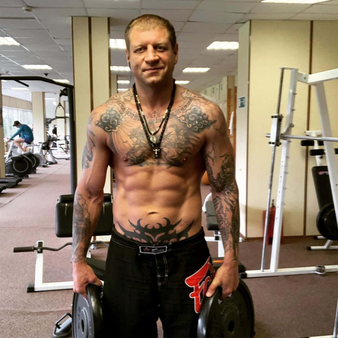 俄羅斯MMA選手亞歷山大·埃米利亞年科在健身房 壁紙 — 一個有紋身的肌肉男赤裸上身站在健身房，自信地微笑著拿著槓片。