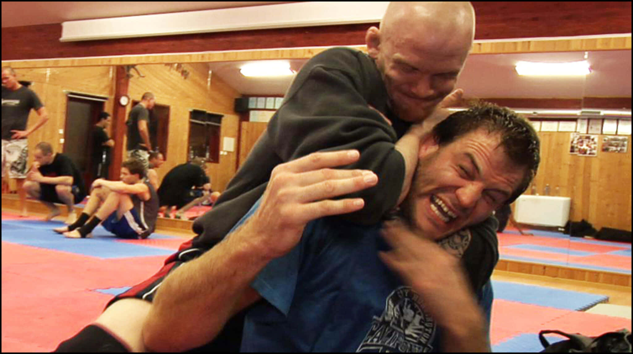 挪威MMA選手約阿希姆·漢森執行後裸頸絞技 壁紙 — 兩名男子在木牆武術道場的藍紅墊上激烈纏鬥，其他人在旁觀看。