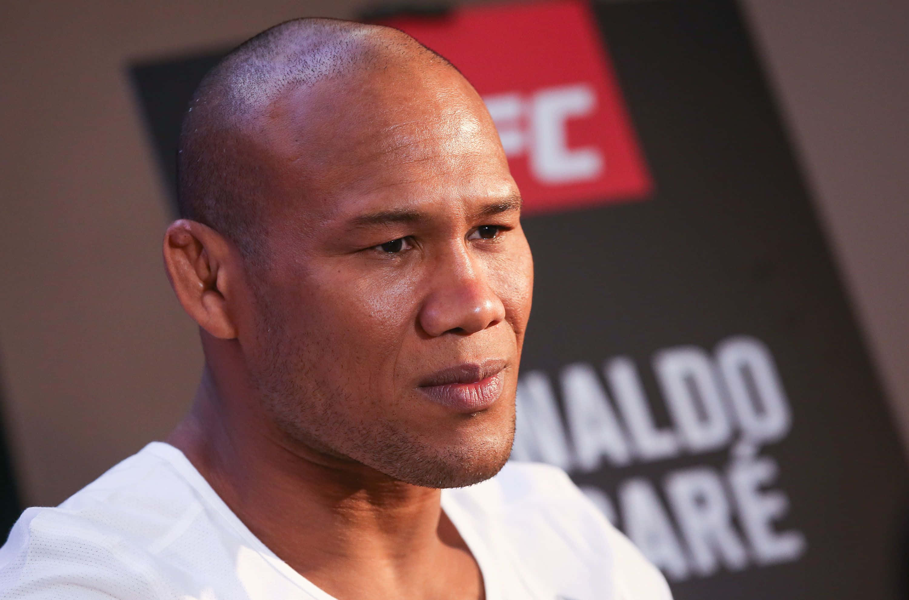 巴西知名MMA選手羅納爾多·蘇薩的特寫鏡頭 壁紙 — UFC記者會上，一名光頭男子身穿白恤衫的close-up肖像，背景品牌標誌模糊。