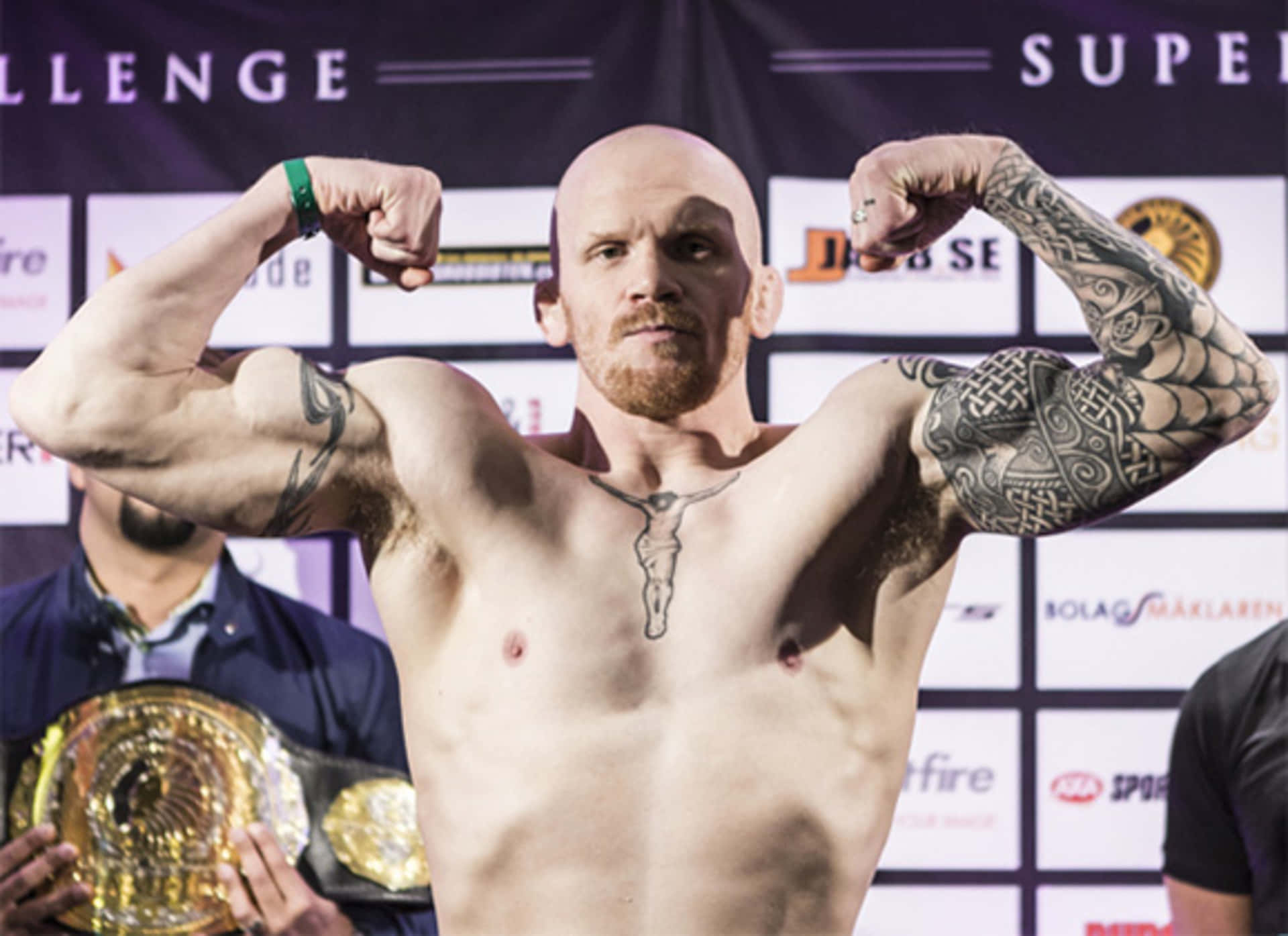 挪威MMA選手約阿希姆·漢森於Superior Challenge 11主賽稱重儀式展現肌肉 壁紙 — 一位肌肉發達的光頭拳手，身上有紋身，在稱重活動中戴著冠軍腰帶，彎曲手臂。