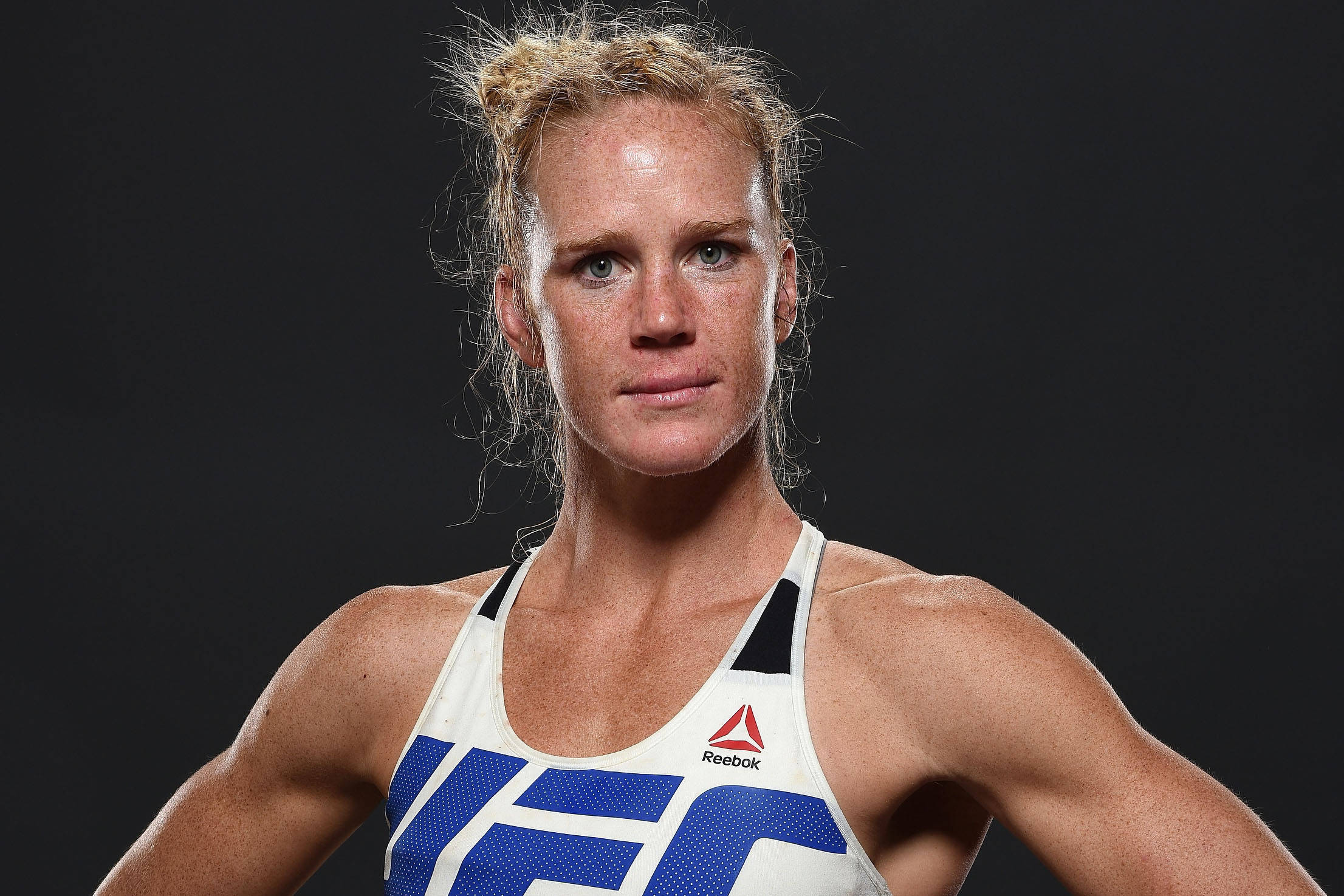 MMA傳奇Holly Holm 壁紙 — 一位堅定的女性MMA拳手，身穿白色和藍色的Reebok UFC上衣，在深色背景下凝視著。