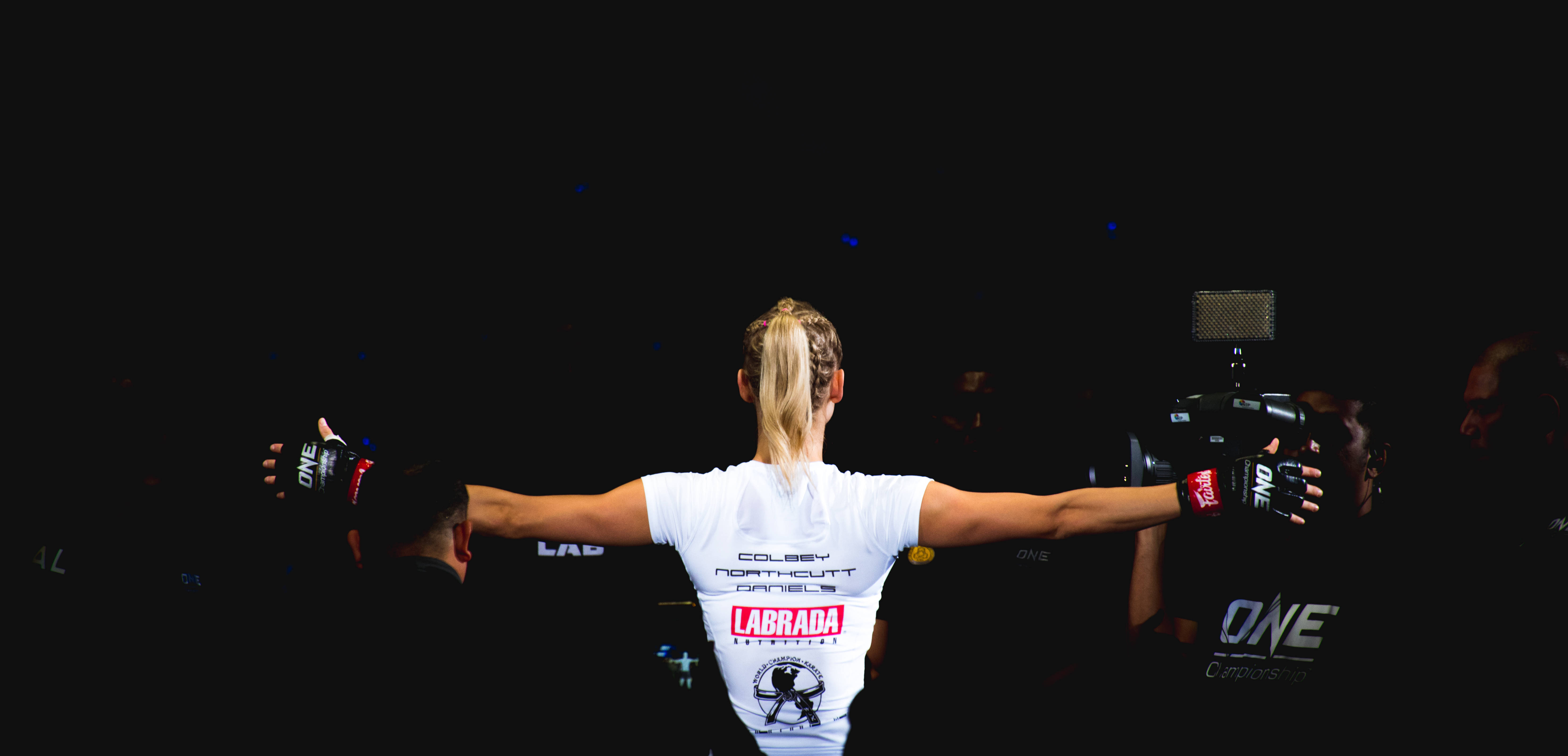 MMA選手Colbey Northcutt 壁紙 — 一名女戰士在黑暗的競技場中張開雙臂，背對鏡頭，在明亮的聚光燈下慶祝勝利。