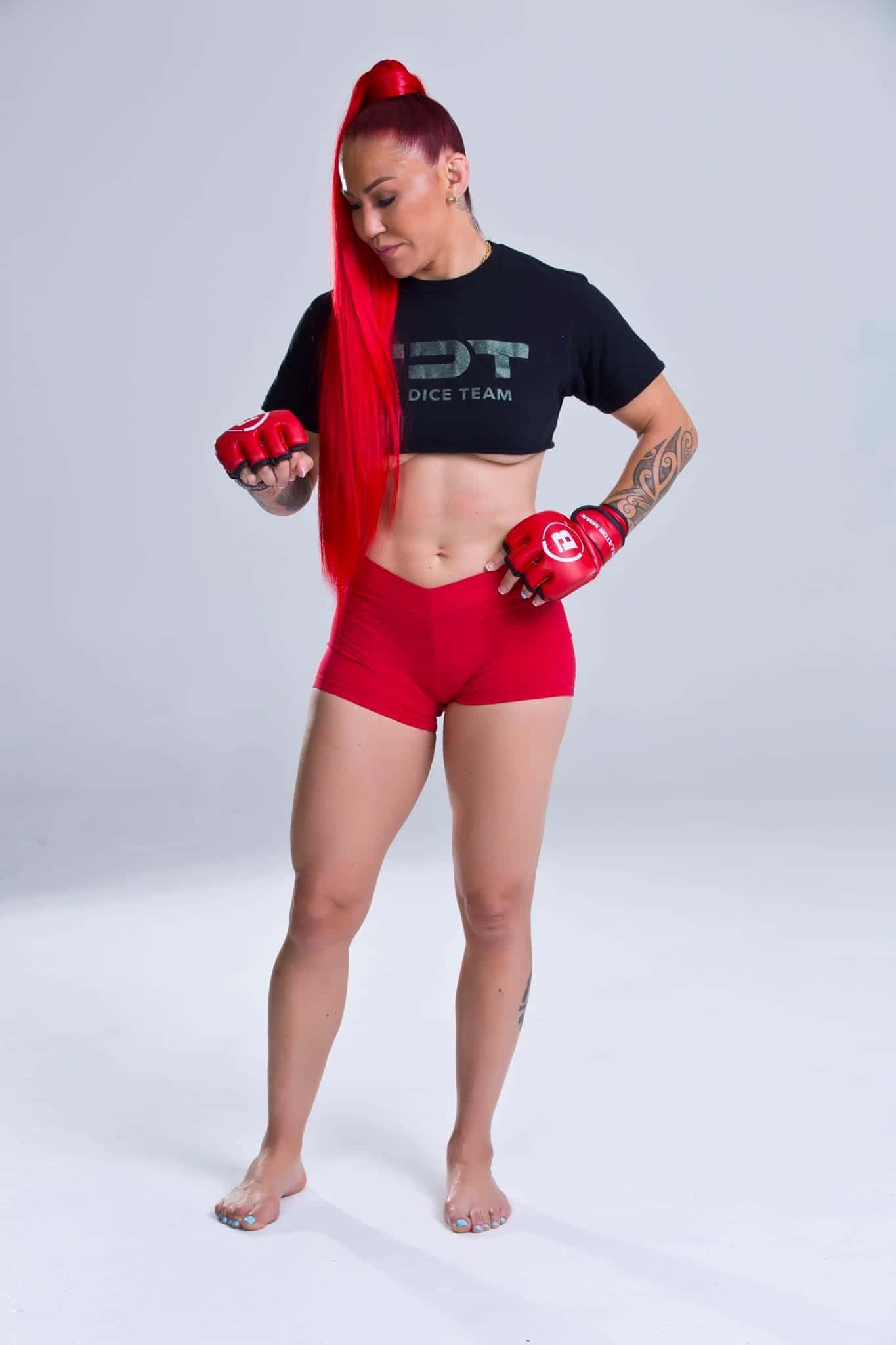 MMA選手Cris Cyborg全身寫真照 壁紙 — 一位擁有鮮豔紅髮和手套的女戰士，在工作室中自信地擺姿勢，背景是純白色。
