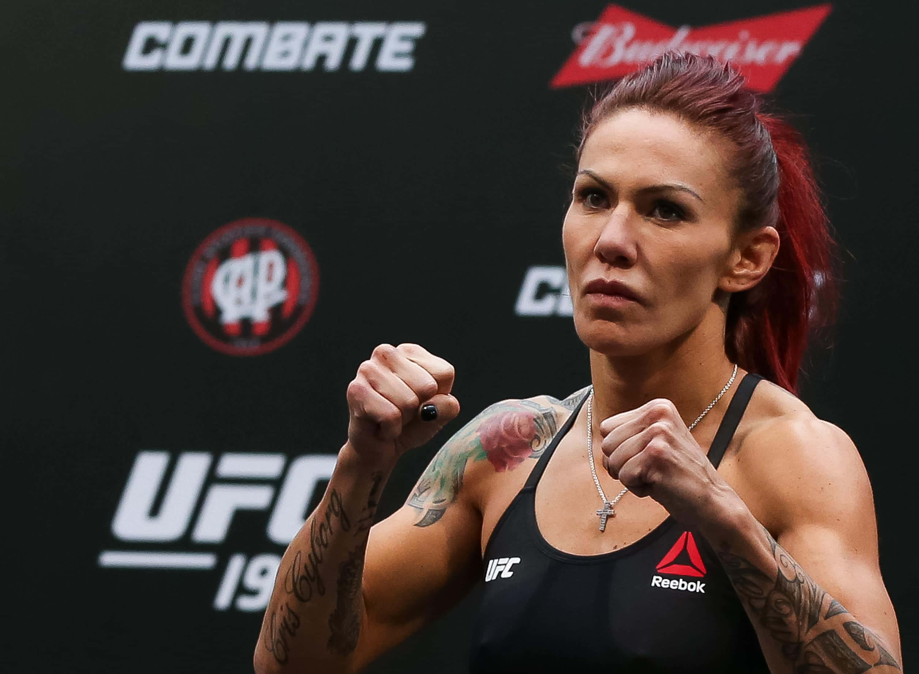 MMA選手Cris Cyborg緊緻特寫照 壁紙 — 一位專注的女性MMA拳手，紅髮及紋身，在UFC背景前擺出戰鬥姿勢。