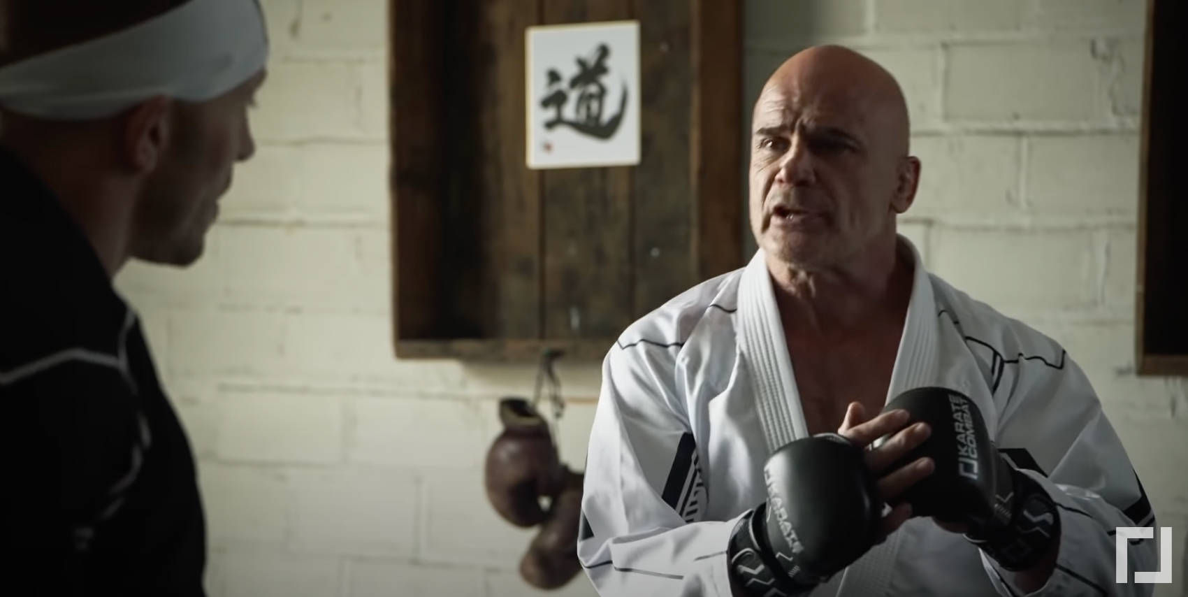MMA選手巴斯·魯滕 壁紙 — 一位嚴肅的光頭武術教練穿著白色道服，在道場裡面對學生，手持黑色手套，牆上掛著「道」字卷軸。