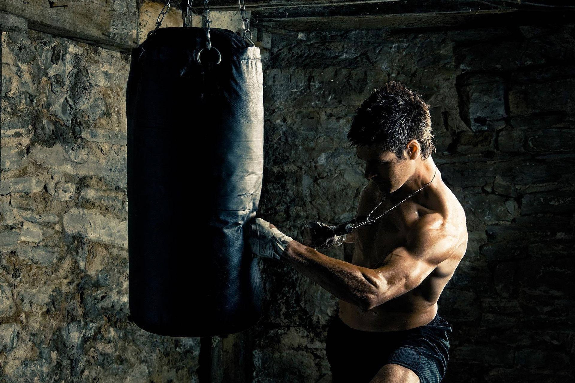 MMA選手拳擊訓練 壁紙 — 一個肌肉發達的男人戴著手套在光線昏暗的石牆健身室中，以戲劇性的燈光重擊沙包。