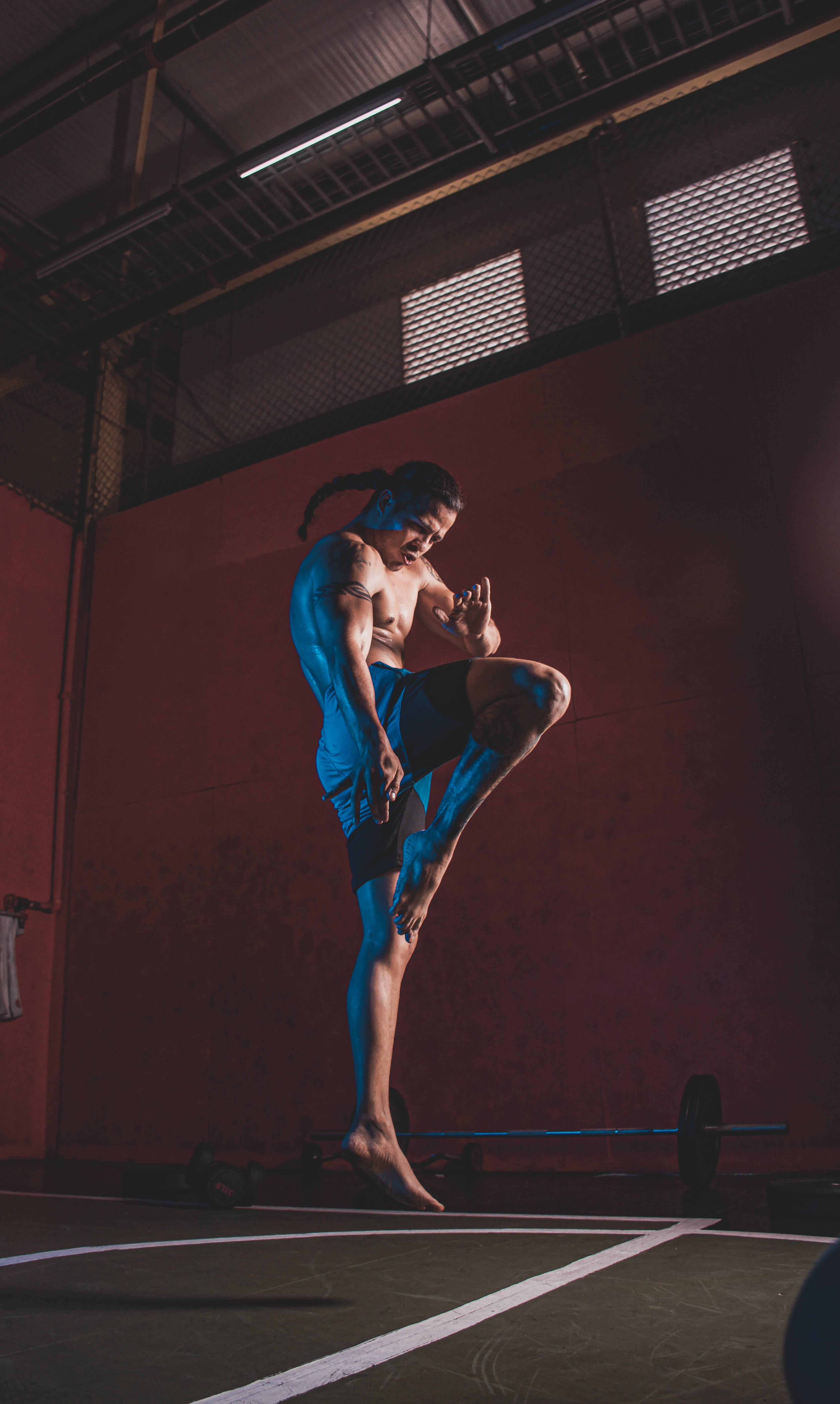 MMA選手獨自訓練 壁紙 — 一位肌肉發達的男子穿著藍色短褲，在光線昏暗、紅牆和裸露天花橫樑的健身房中執行高踢腿動作。