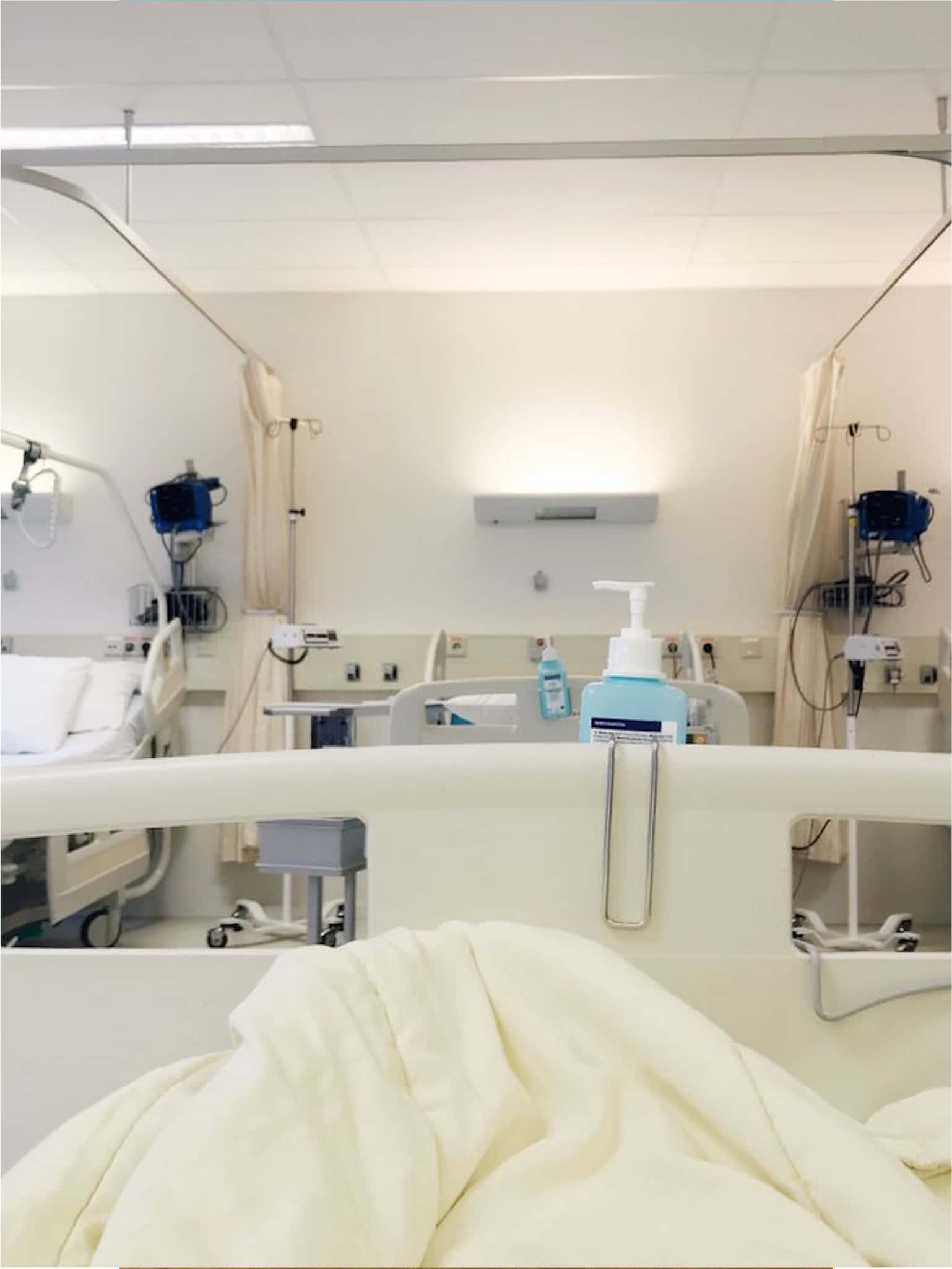 Hospital Bed 桌布 壁紙 — 一個無菌的醫院房間，前景有病人床，背景有醫療設備，在明亮的螢光燈下。