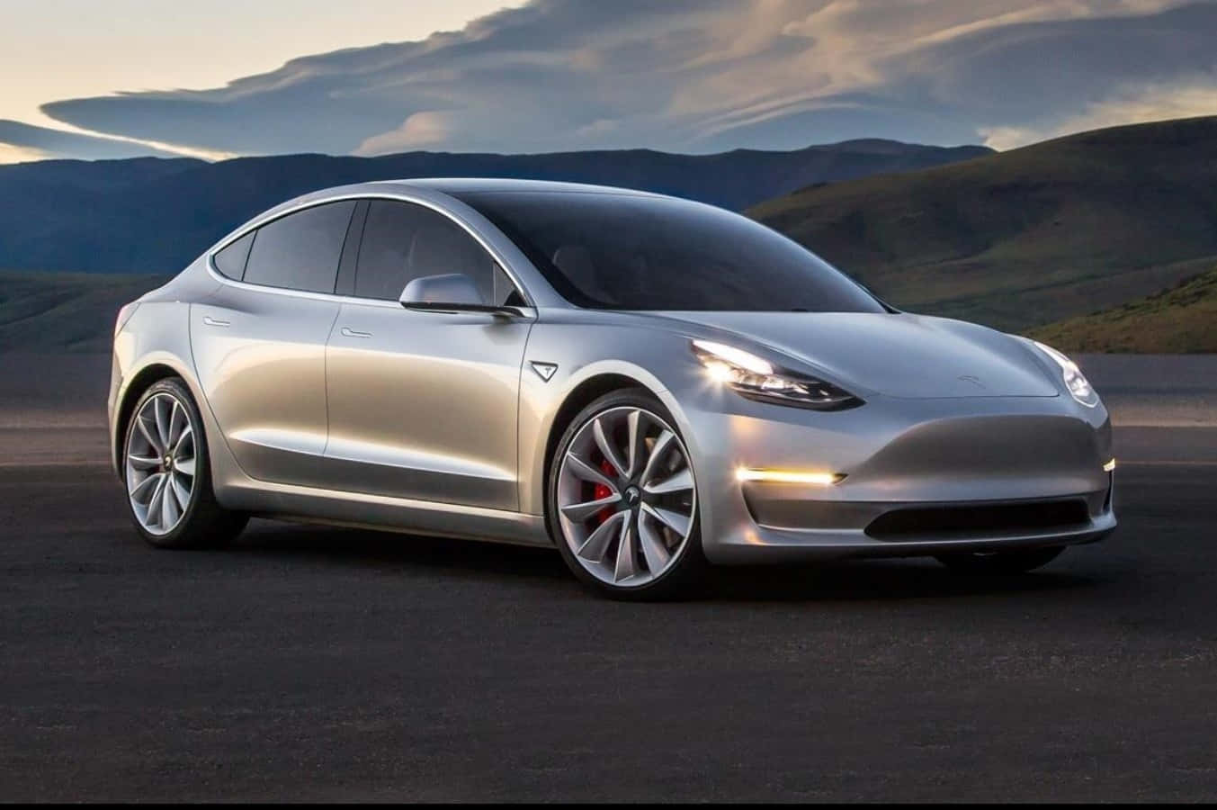 引領電動車未來：特斯拉Model 3 壁紙 — 一架時尚的銀色 Tesla Model 3 停泊在黑暗的道路上，背景有山脈和多雲的天空。