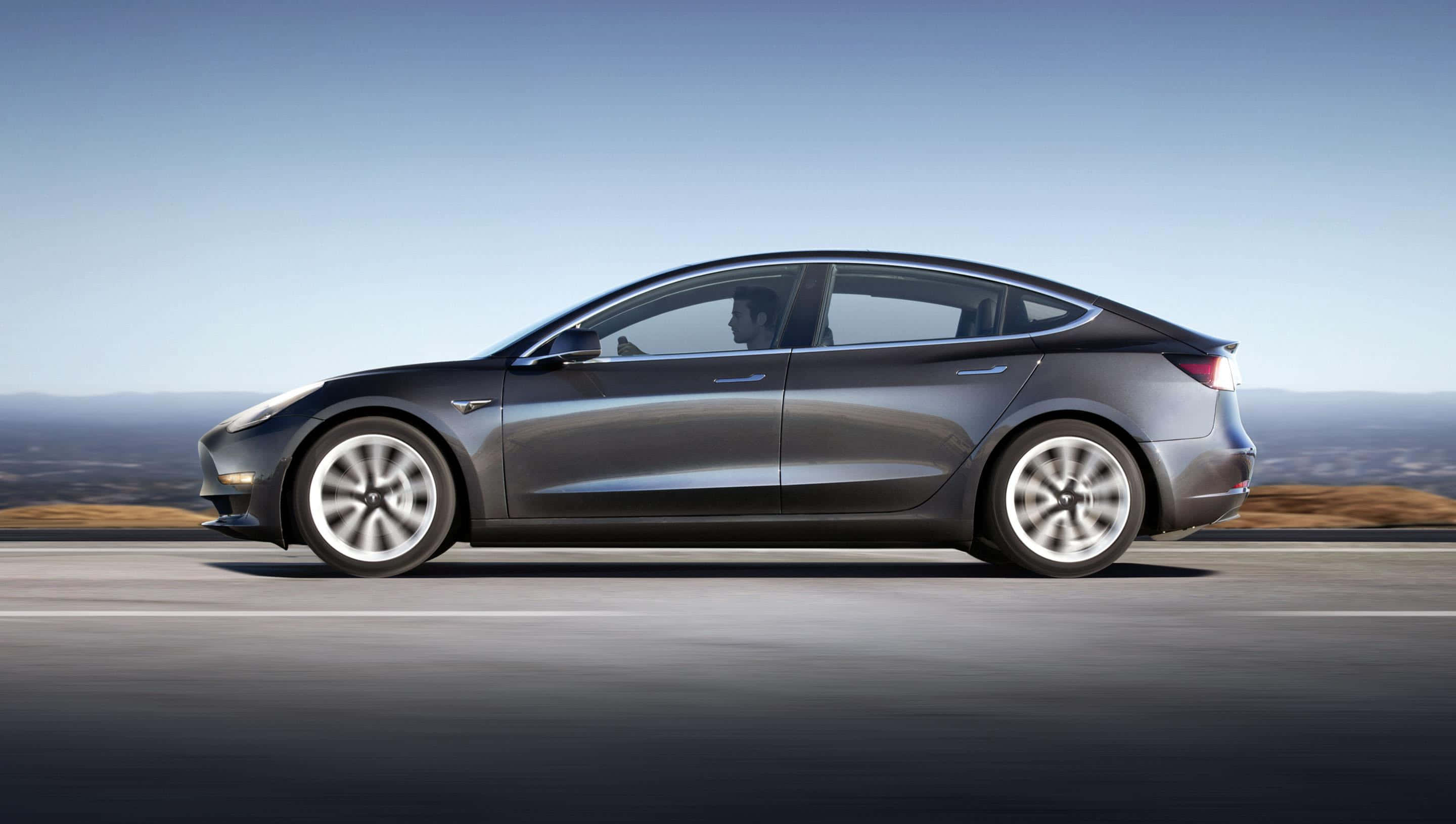 時尚與創新，特斯拉 Model 3 壁紙 — 一架流線型灰色Tesla Model 3在高速公路上行駛，背景是清晰的藍天和遠山，帶有動態模糊效果。