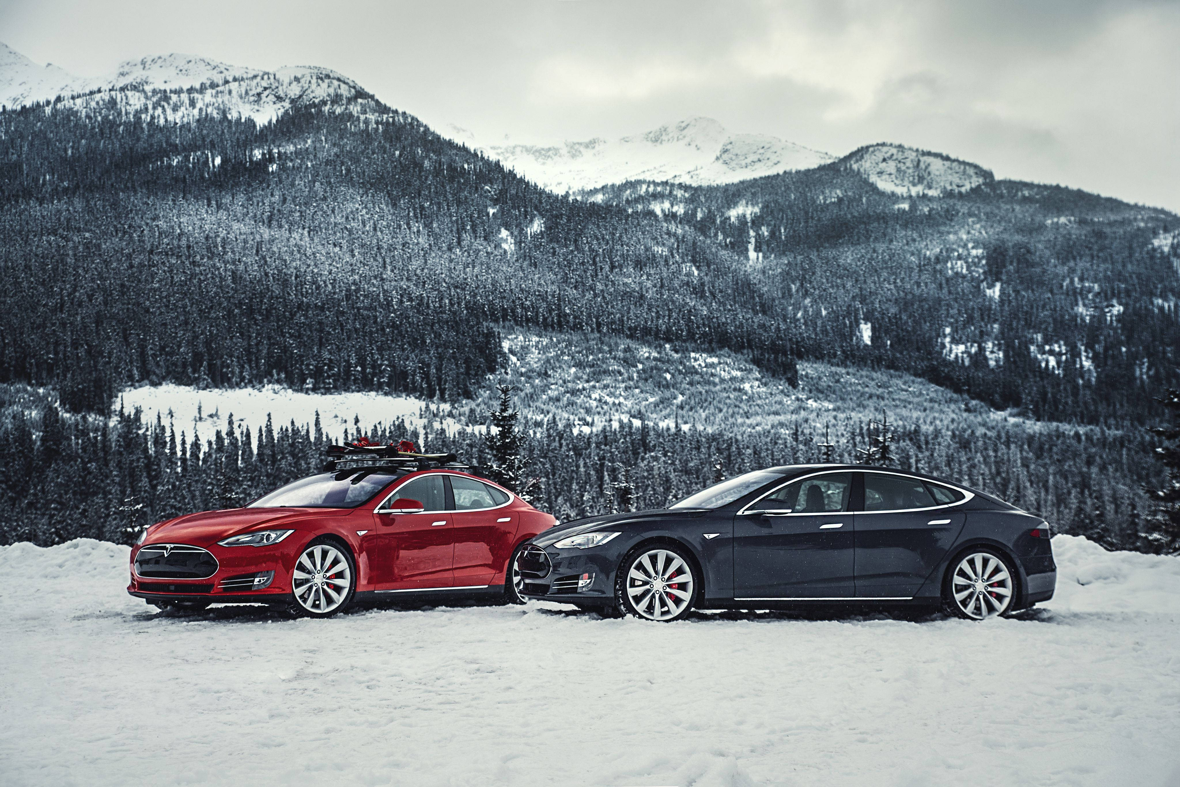 特斯拉Model S P85D紅黑配色 壁紙 — 兩輛紅色和黑色的Tesla汽車並排停放在雪地上，背景是白雪覆蓋的山脈和常綠森林，天空多雲。