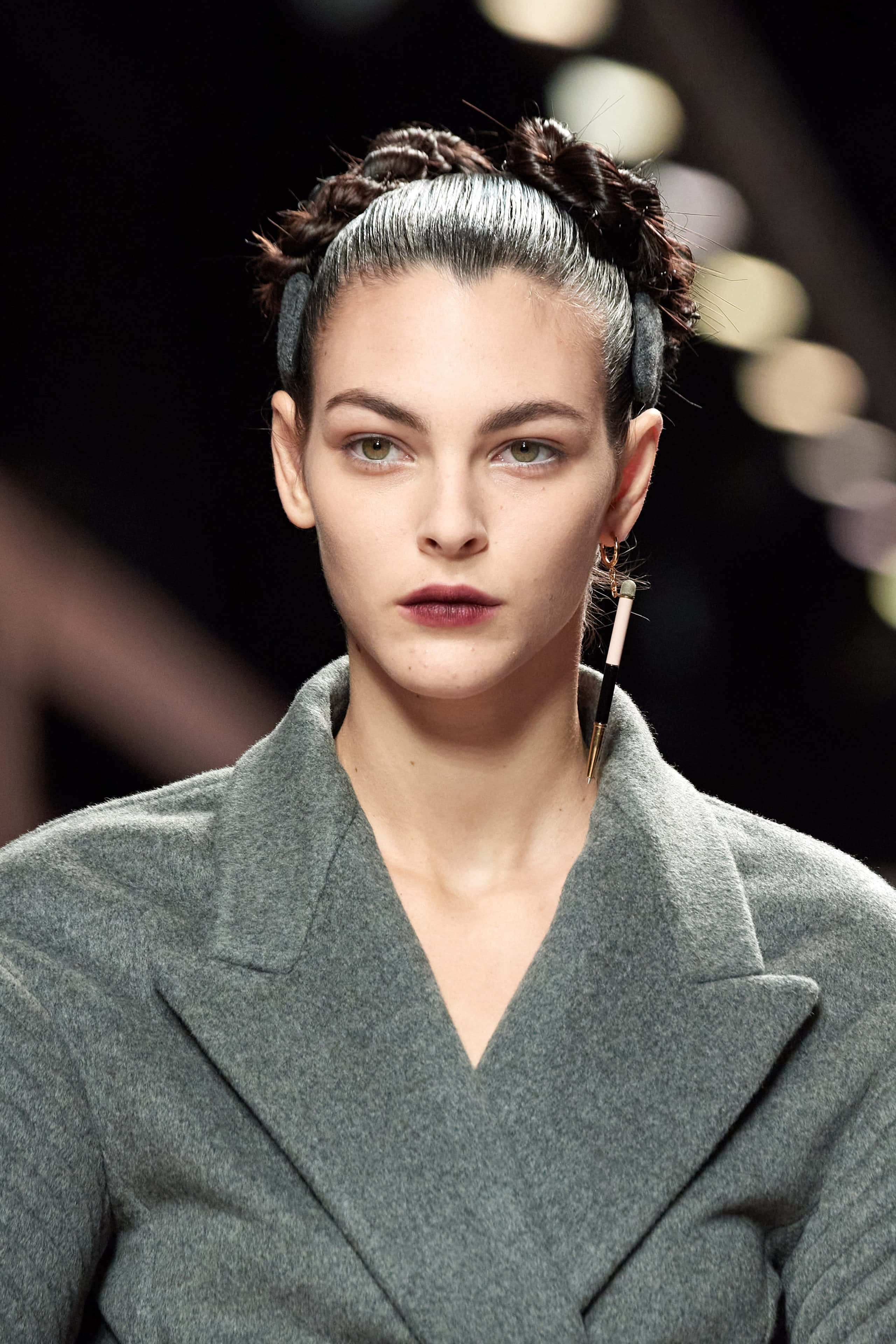 Tiarnie Coupland 桌布 壁紙 — 一位辮子頭髮、大膽妝容的模特兒在 runway 燈光下自信地穿著灰色外套行走。