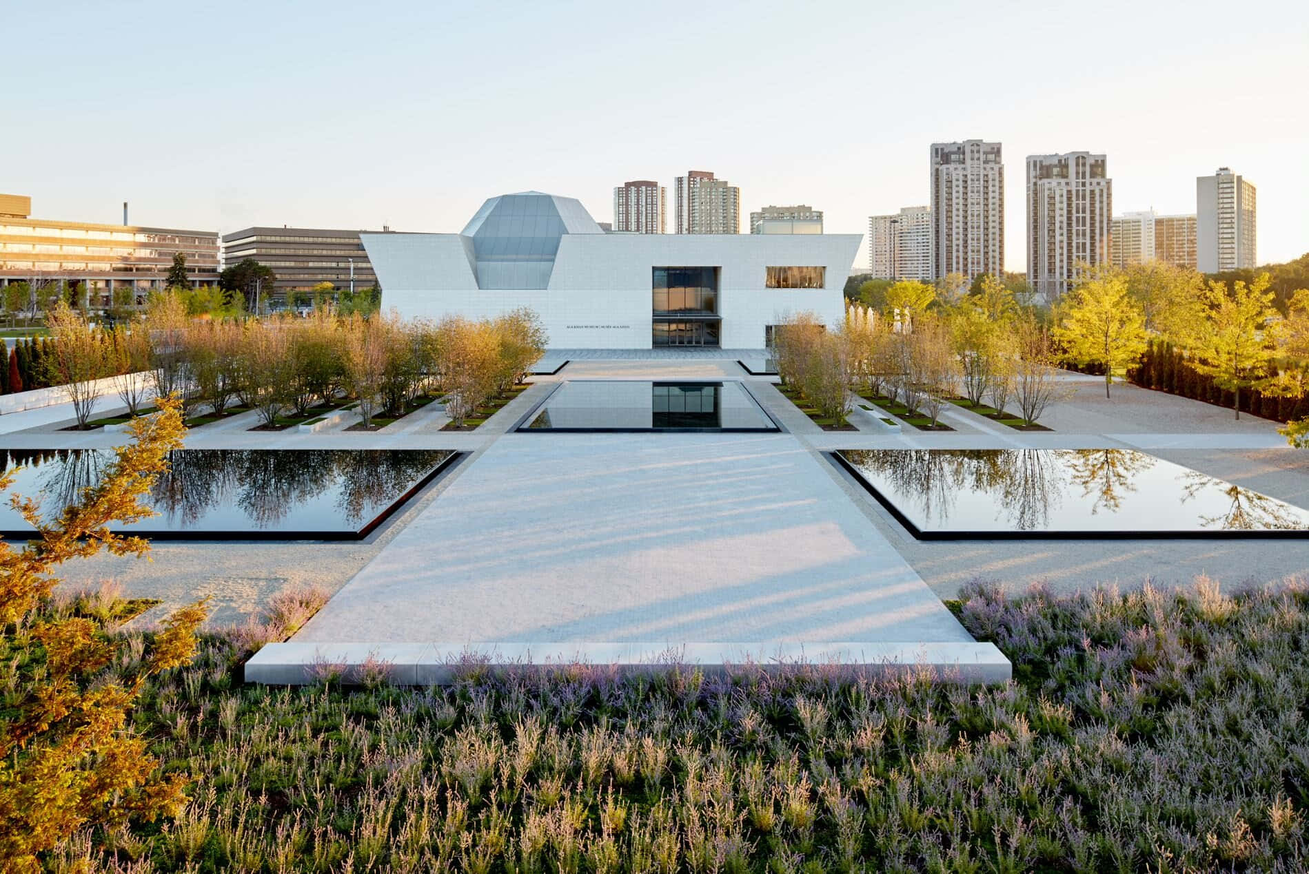 Aga Khan Museum 桌布 壁紙 — 一座現代白色建築，帶有玻璃圓頂，位於長長的倒影池盡頭，周圍環繞著秋樹和紫色草。
