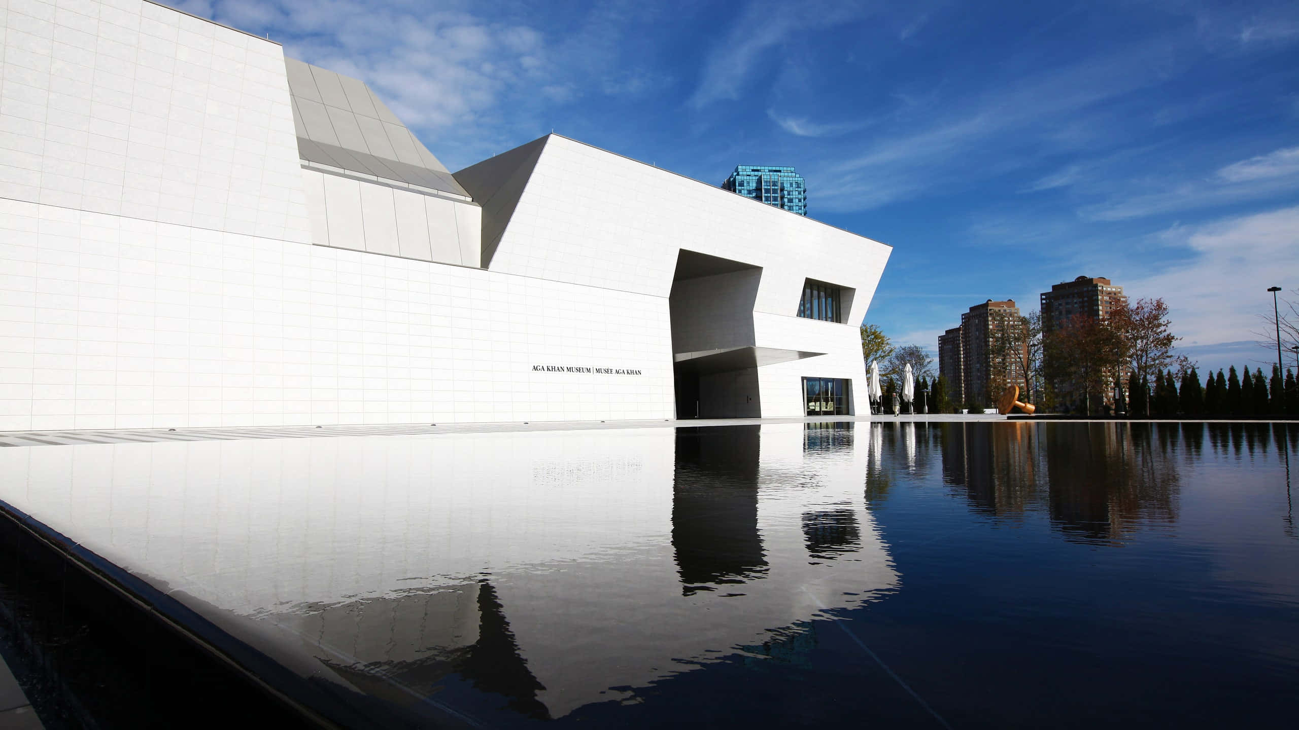 Aga Khan Museum 桌布 壁紙 — 一座現代白色幾何博物館建築在晴朗藍天白雲下，完美倒映在平靜的水池中。