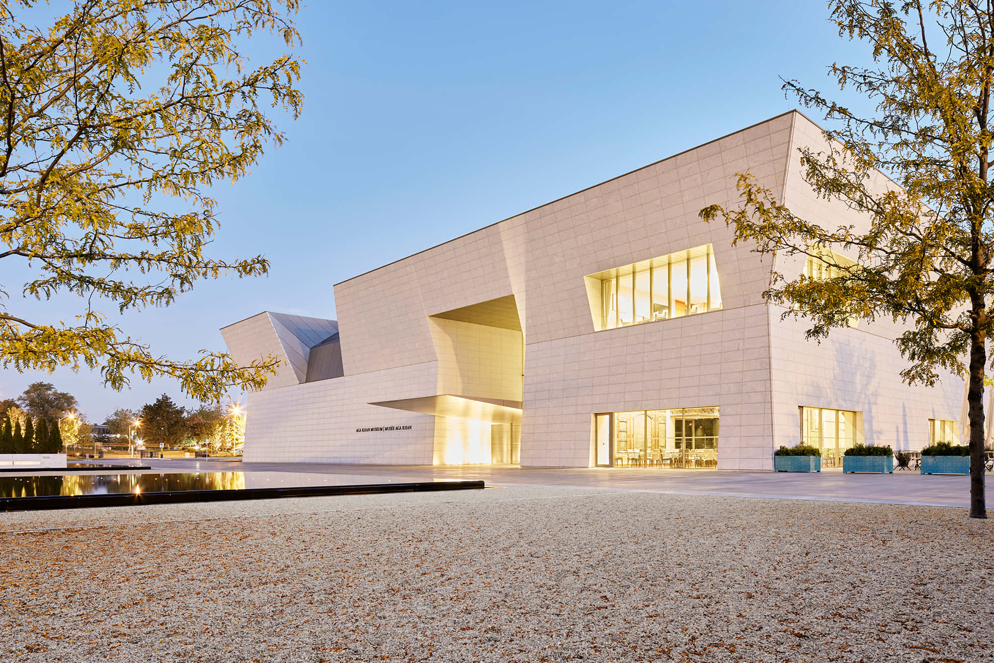 Aga Khan Museum 桌布 壁紙 — 現代白色博物館建築，在黃昏時分被秋樹環繞，窗戶發光。